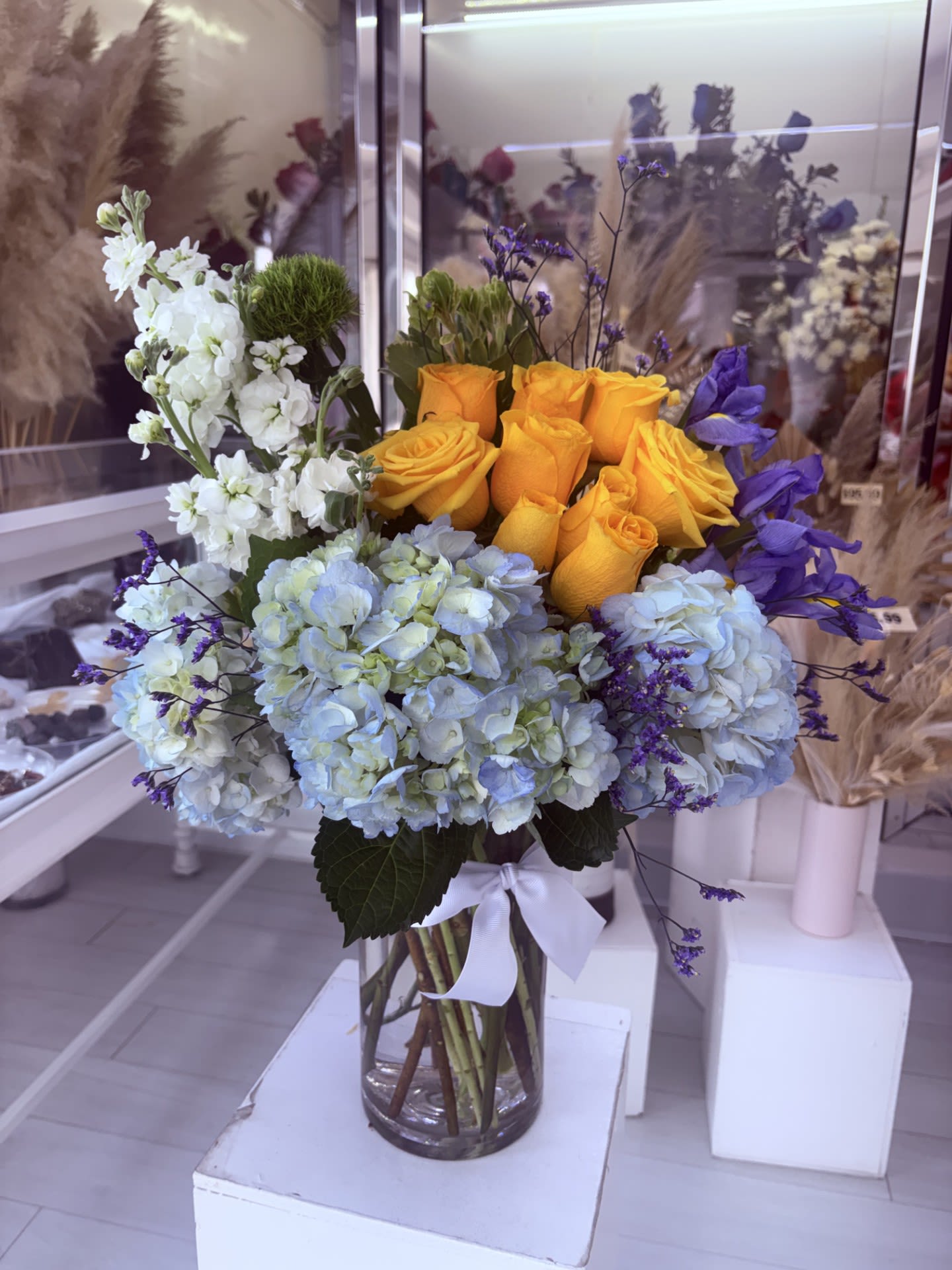 Vase arrangement of yellow roses, blue hydrangeas, white blooms and purple accents in a clear glass cylinder