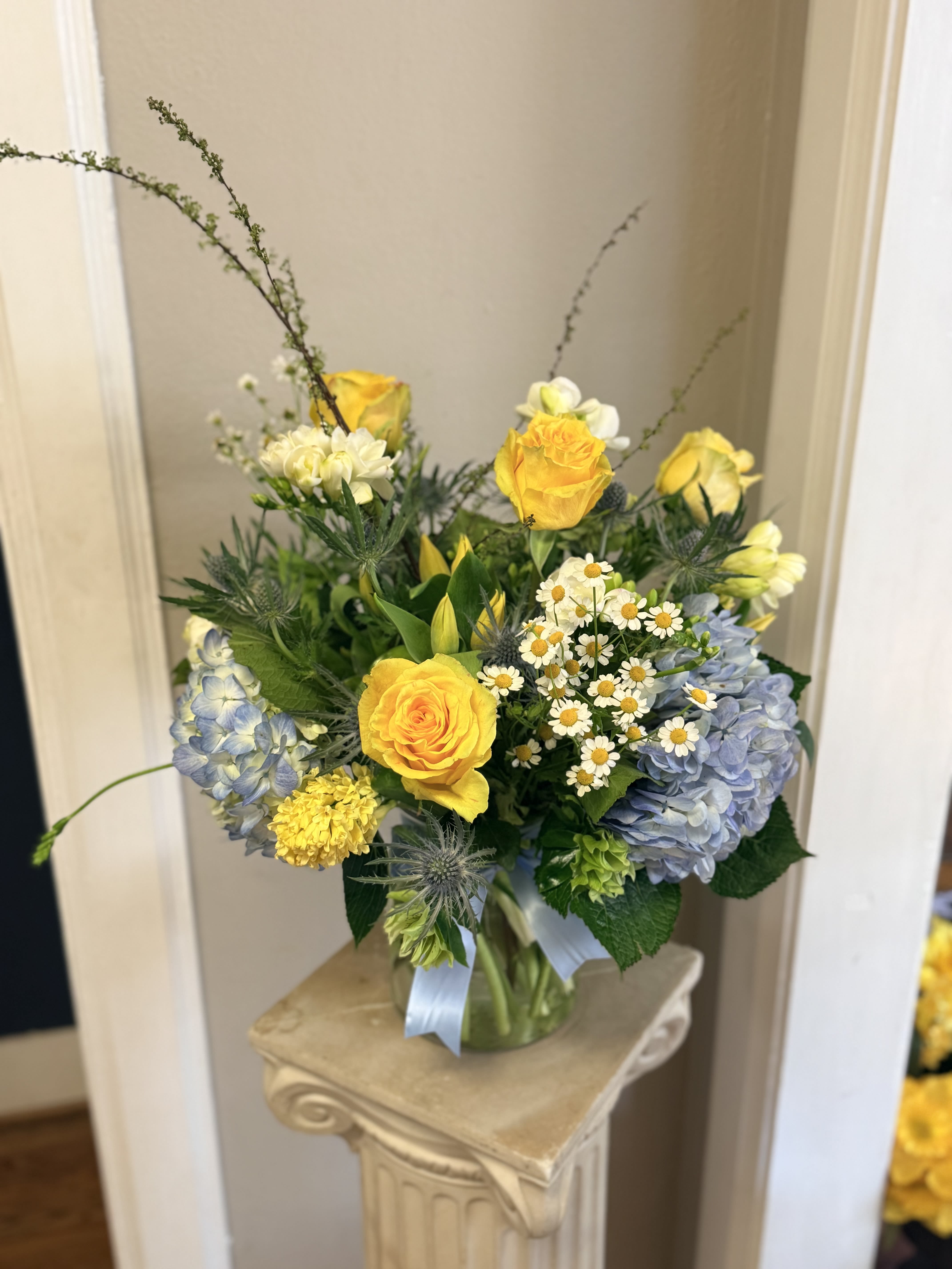 Cottage Hill  - Sunlit yellow roses headline this airy flower arrangement, the perfect birthday bouquet or spring thank-you gift that feels effortlessly cheerful. Pale blue hydrangeas and small white daisy-like blooms soften the composition, creating a fresh, uplifting look your recipient will notice the moment it arrives. Its asymmetrical, garden-inspired shape reads as casual and thoughtful-ideal for saying "congrats," "thank you," or "just because" in a truly personal way.  The Cottage Hill arrangement is built in a clear glass vase tied with a light blue ribbon for a polished, gift-ready finish. Tall branching accents rise above a fuller base of mixed roses, hydrangeas, daisies and textured greenery, giving pleasing height and a loose, natural silhouette. Colors are sunny yellow, pale blue, white and verdant green for a springtime palette that remains wearable any time of year.  Send this to someone who deserves to feel seen and celebrated-whether it's a birthday, a job milestone, or a spontaneous pick-me-up. The vase presentation makes it easy to display right away; for best longevity, refresh the water every two days and trim stems slightly when re-cutting to keep blooms vibrant.