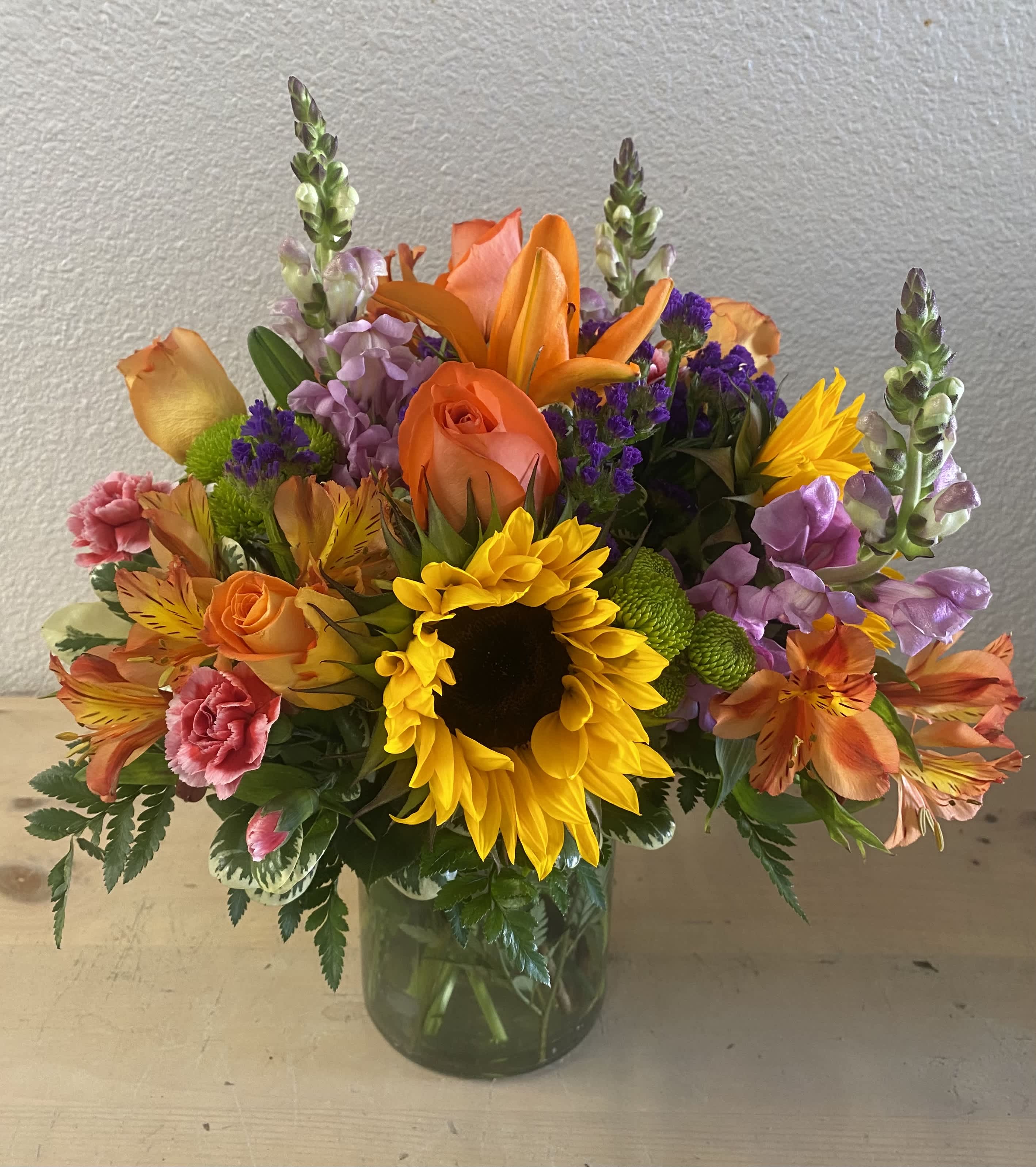 Colorful bouquet with sunflowers, roses, lilies, and purple filler in a glass vase