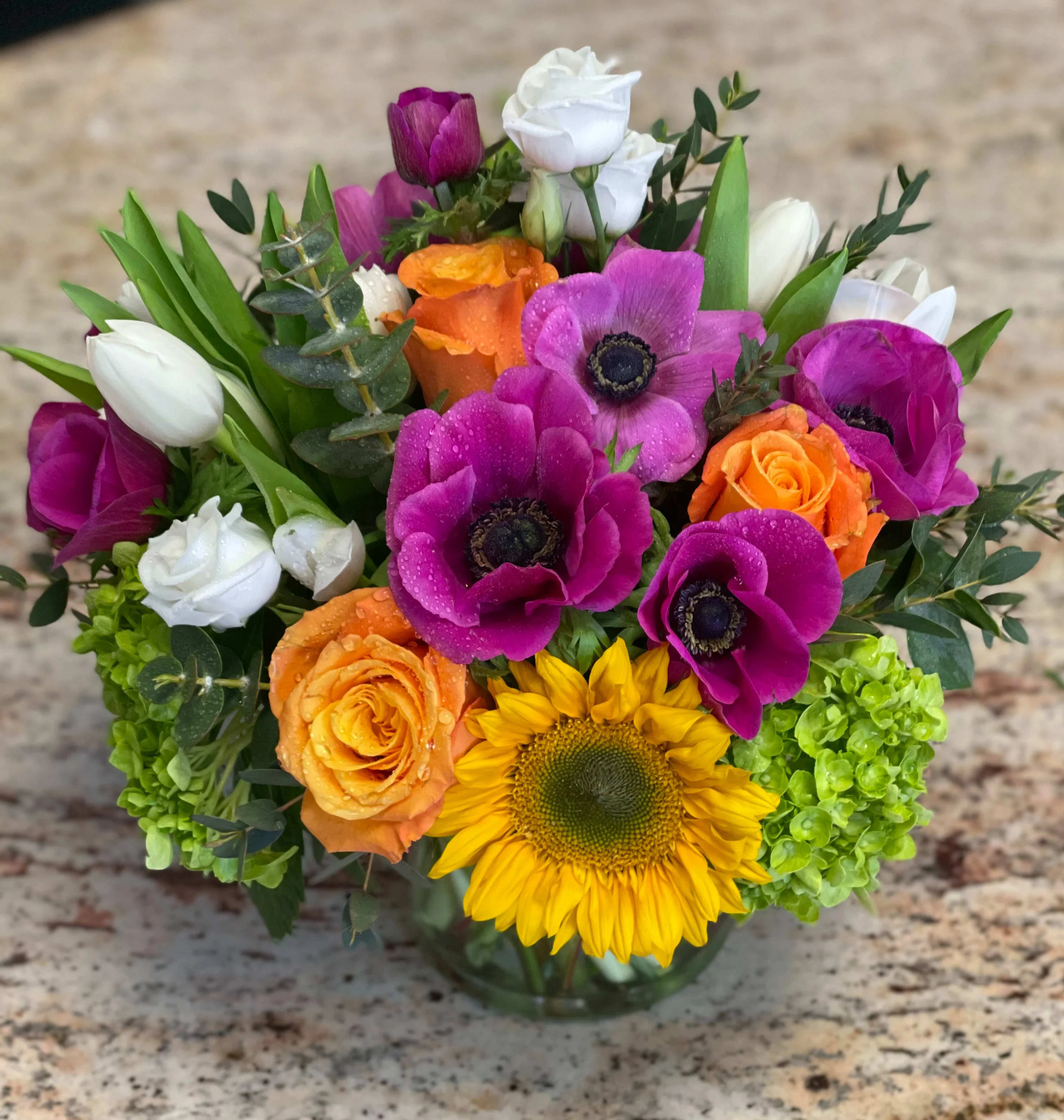 Colorful bouquet with sunflowers, roses, tulips, and white blooms in a glass vase
