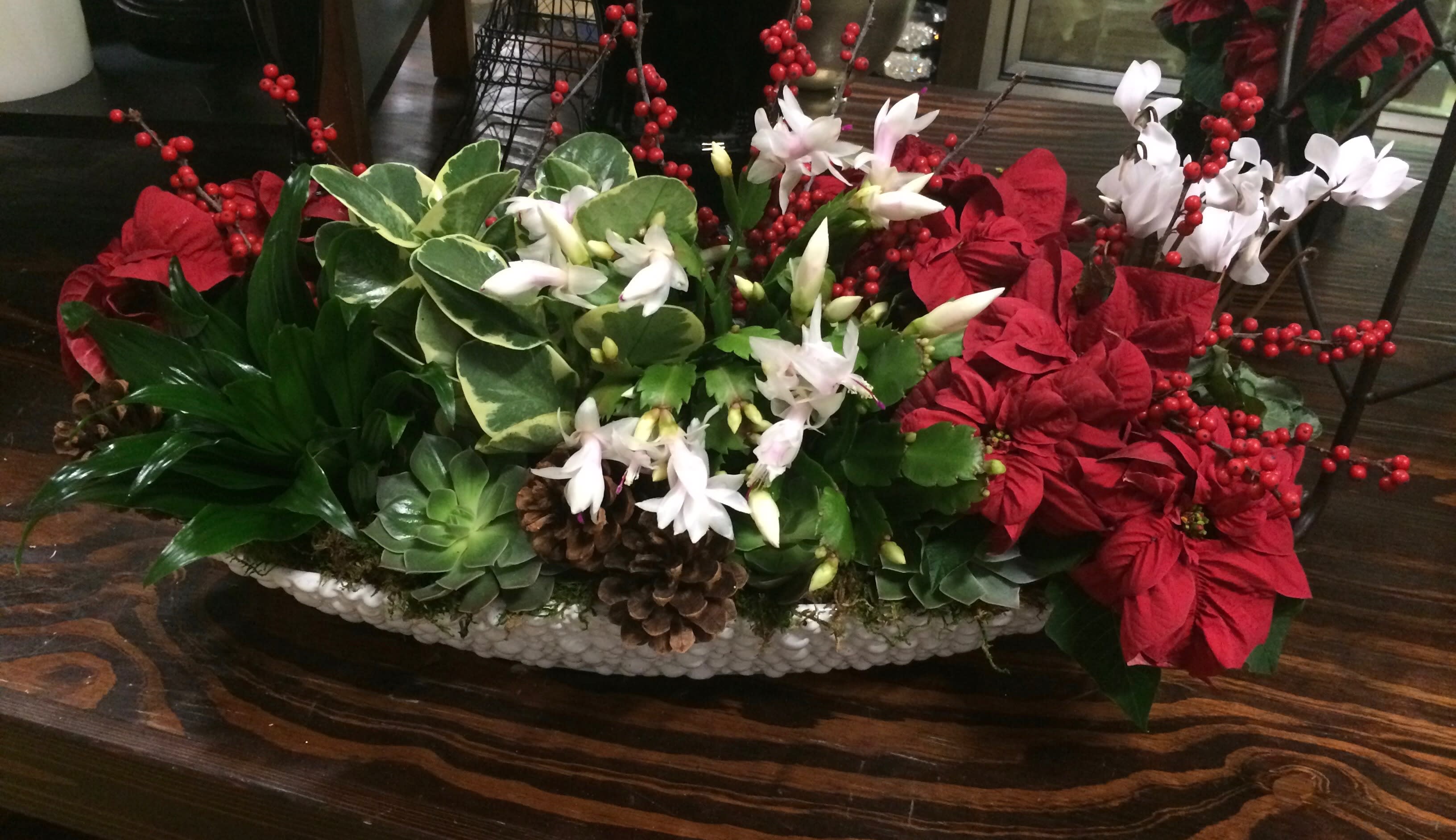 Holiday centerpiece with red poinsettias, white flowers, and pinecones in a low white container