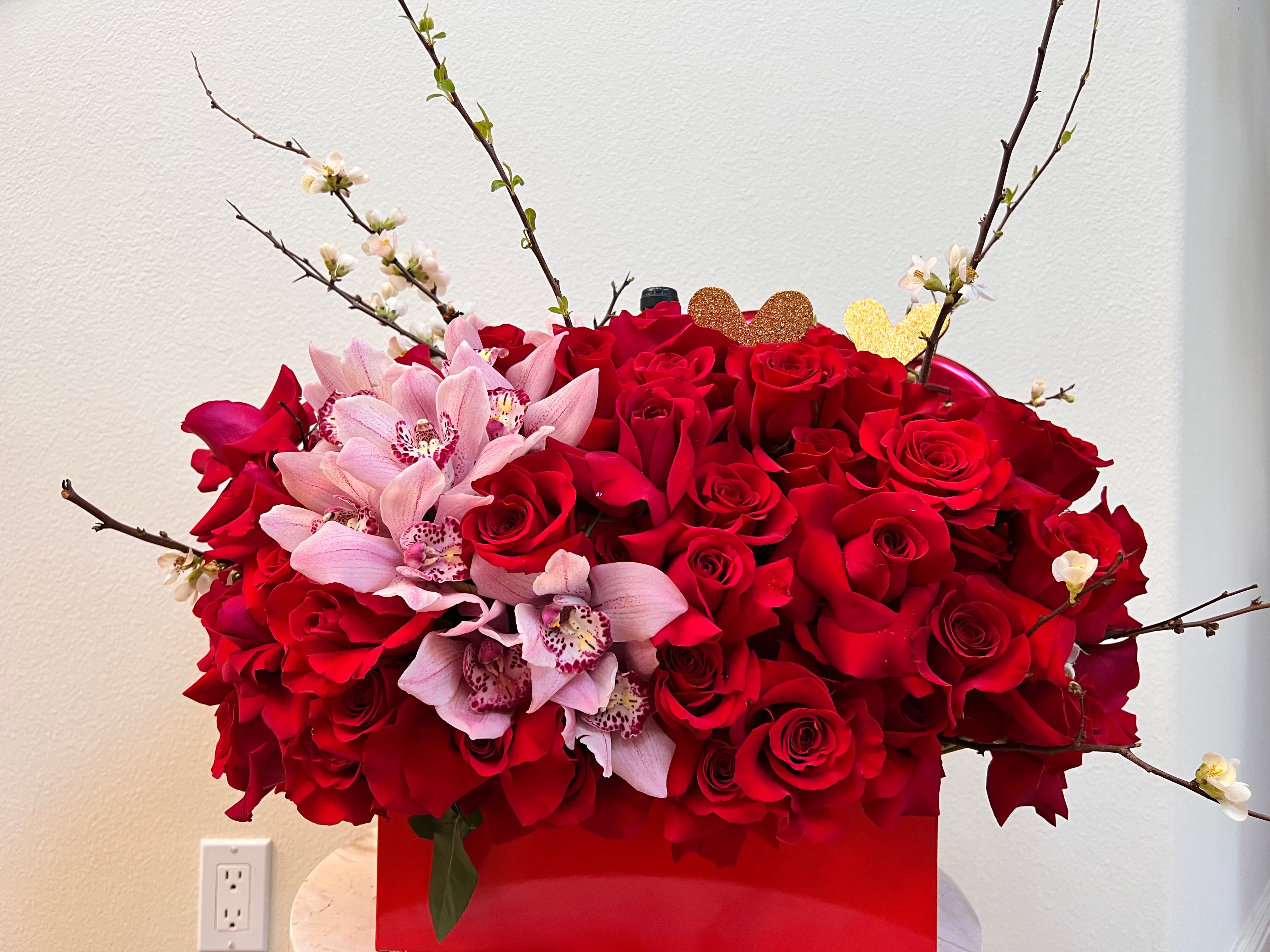 Red roses with pink orchids in a red box, accented by flowering branches.