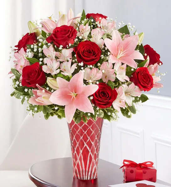 Red roses and pink lilies in a red glass vase beside a small gift box