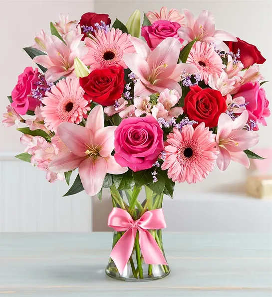 Pink lilies, roses, and gerbera daisies in a glass vase with a pink ribbon