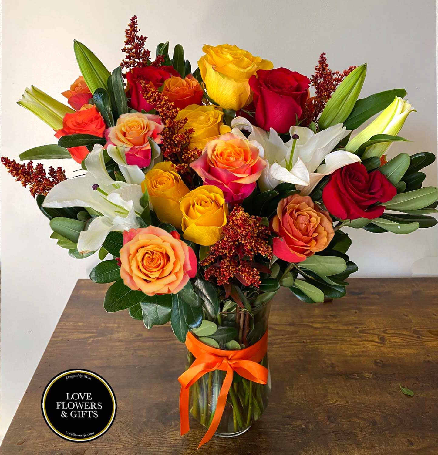 Tall vase arrangement of red, yellow, and orange roses with white lilies and an orange ribbon bow.