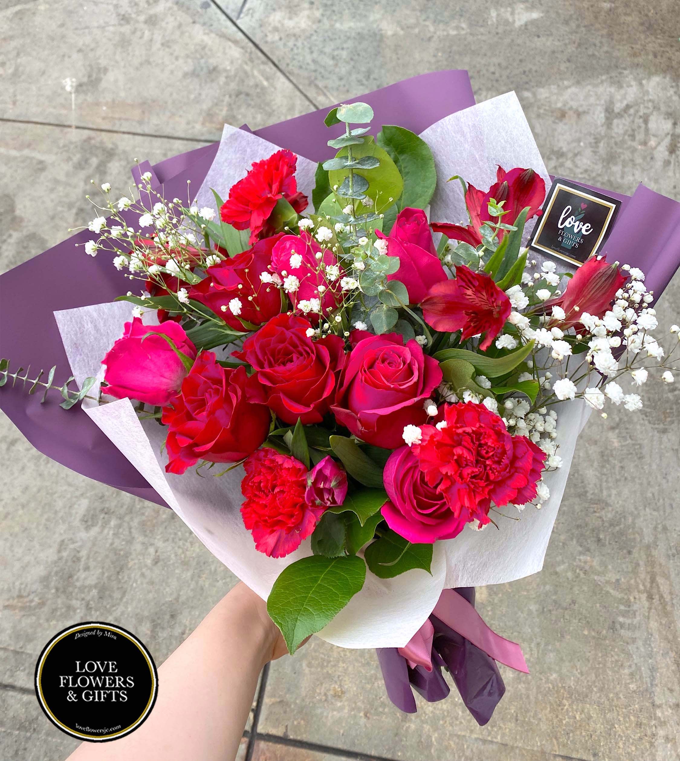 Hand-tied bouquet of red roses, carnations, and white filler flowers wrapped in purple and white paper.