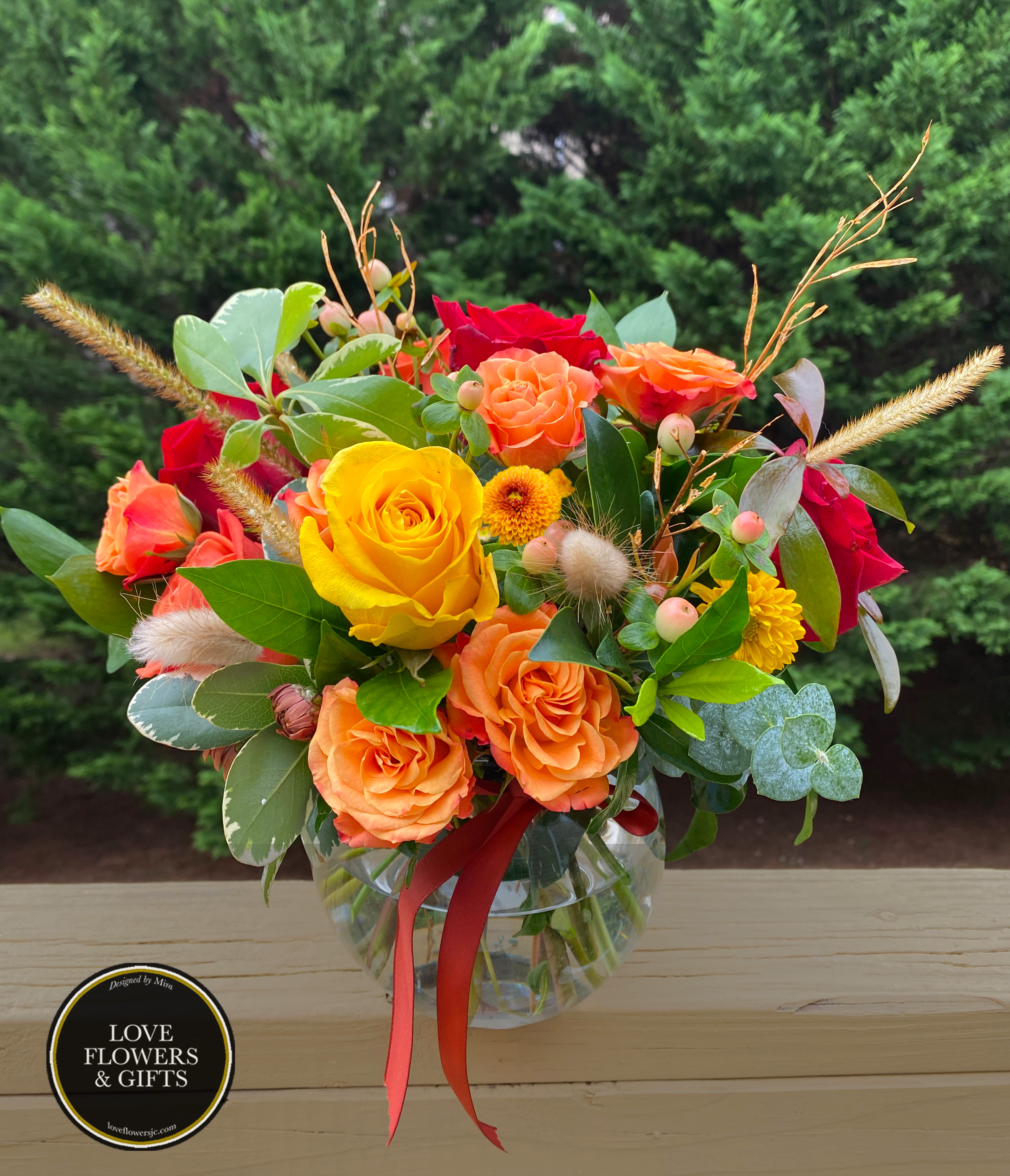 Round glass vase of yellow, orange, and red roses with small yellow mums and berries, tied with an orange ribbon