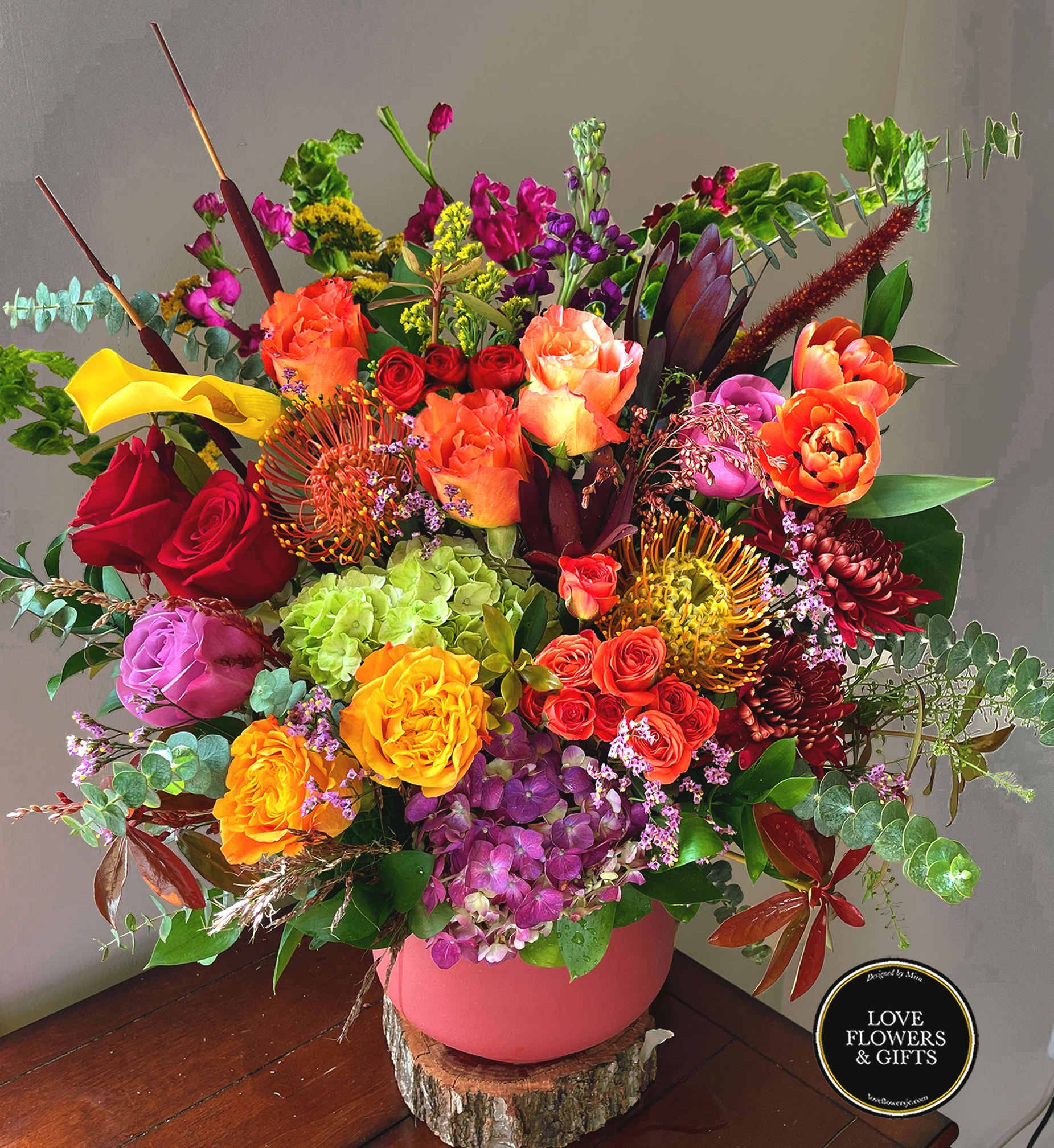 Large colorful arrangement of roses, hydrangeas, protea and mixed blooms in a pink vase on a wood slice