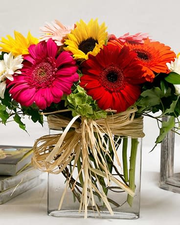 Bright gerbera daisies arranged in a clear square vase with raffia ribbon