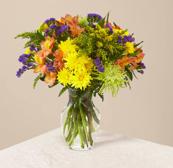 Mixed bouquet of yellow and green mums, orange alstroemeria, and purple flowers in a clear glass vase