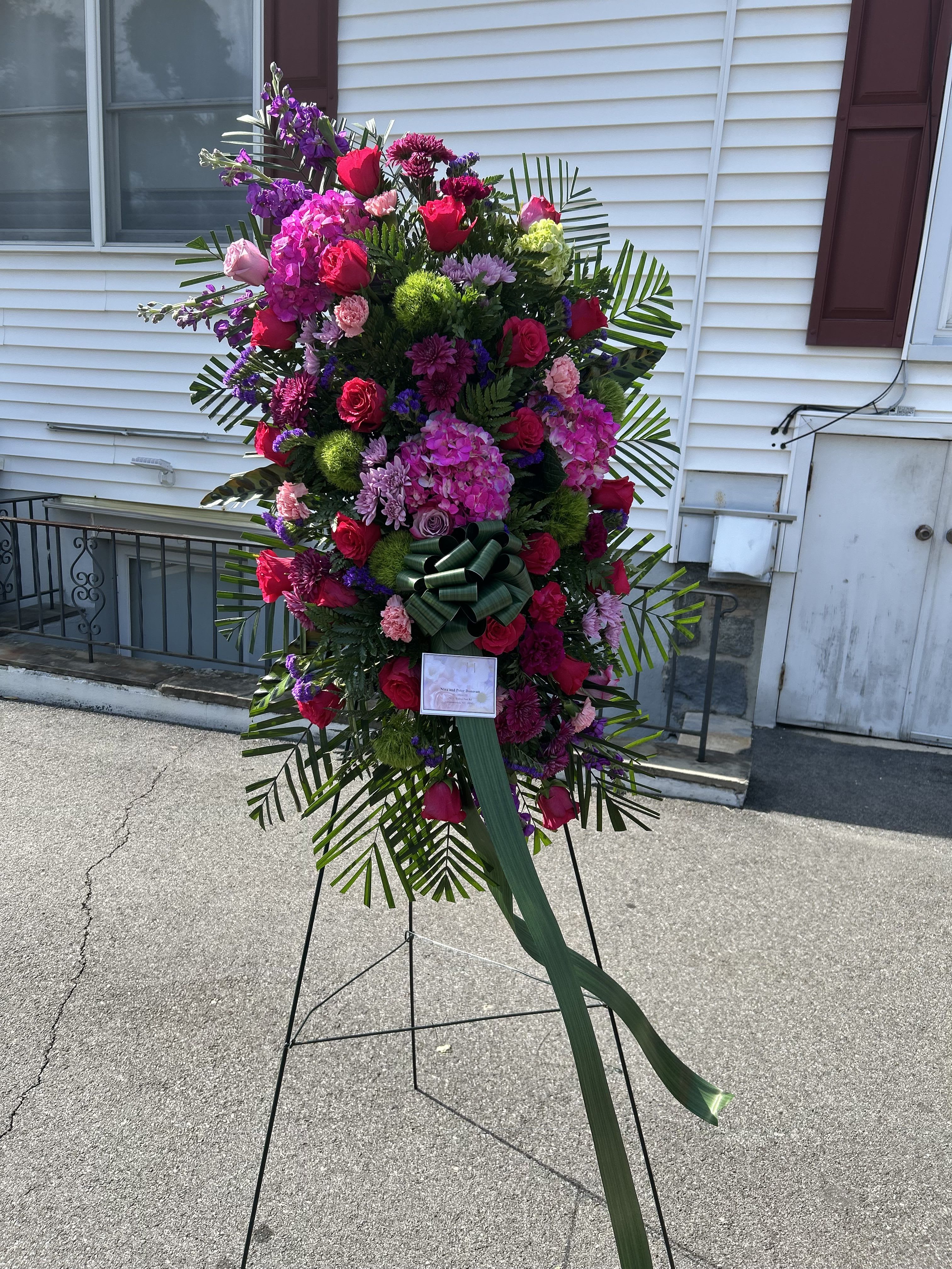 Refreshing Spray in Pink - A dramatic standing spray in pink, purple, red and green that makes a solemn, heartfelt statement for sympathy or memorial services. This standing spray for sympathy places a dense, rounded center of roses, carnations, chrysanthemums and hydrangea clusters where the eye naturally rests, with alstroemeria and smaller accent blooms adding delicate texture. The tall, formal silhouette and palm-like greenery create a ceremonial presence that helps the person you're honoring feel remembered and respected.  The arrangement sits on a sturdy standing easel and is finished with a prominent green ribbon bow and a floral card for your personal message. Long trailing leaves frame the composition and add graceful movement, while the mix of bloom sizes-from full hydrangea clusters to refined rose heads-gives visual depth and a comforting, layered look. Colors range from soft pinks to richer purples and reds against lush green foliage for a warm, reverent palette.  Perfect for funeral services, graveside tributes, or a memorial celebration, this spray conveys sympathy and support in a dignified way. Include your message on the included card at checkout; the easel and bow are ready for immediate placement, and our florists will arrange blooms to present a fresh, long-lasting tribute on the day of the service.