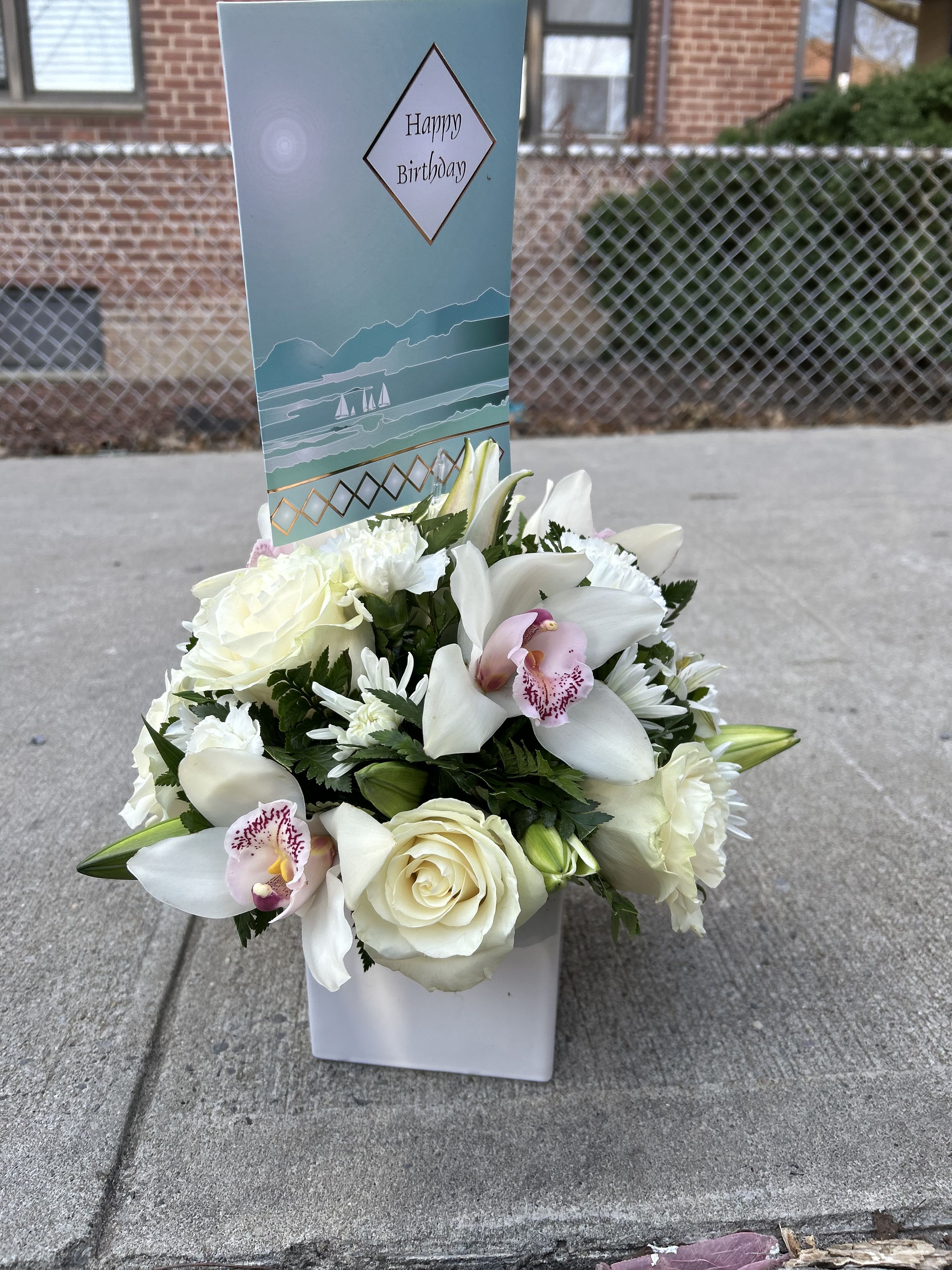 Purity in Square - A clean, airy cluster of white roses, lilies, orchids, daisies and carnations makes this birthday bouquet feel quietly celebratory - perfect as a birthday arrangement or a cheerful everyday gift. The compact square presentation in a white ceramic vase keeps the look modern and refined while soft pink accents in the orchid centers and touches of green foliage add just enough warmth to brighten someone's day. The included tall birthday card behind the flowers gives the person you're thinking of a personalized moment before they even read your message.  White roses and lilies provide the classic fragrance and sculpted form, orchids lend delicate texture and pink-tinged centers, and small daisy-like blooms and carnations fill the arrangement for a soft, full silhouette. Set in a sturdy white ceramic vase, the design sits neatly on tabletops or mantels without needing extra trimming - dimensions suit a compact square display that reads fresh from every angle. This is the kind of bouquet that makes the recipient feel noticed and celebrated; order with your message on the birthday card and we'll prepare it ready to present.