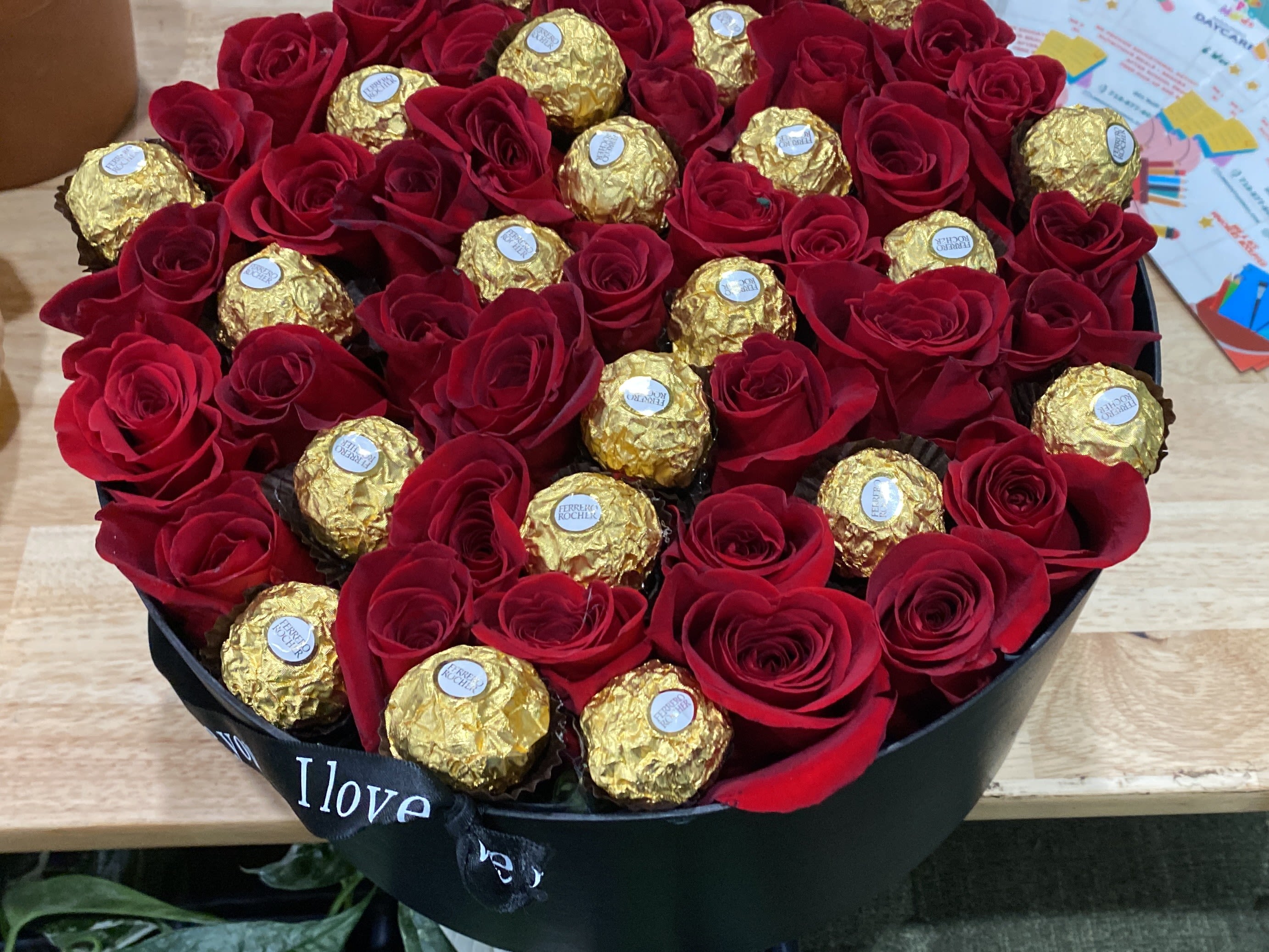 I Love You Box (With Chocolates) - A compact cascade of tightly packed red roses and gold-wrapped chocolates-this flower arrangement for Valentine's Day or an anniversary reads like a bold declaration: "I love you." The red roses give a classic romantic intensity while the metallic gold chocolates add celebratory sparkle; the black floral wrap and ribbon frame the bouquet with dramatic contrast so the person you're thinking of will feel unmistakably treasured.  This arrangement is built as an abundant, gift-ready bouquet rather than a vase piece: deep red roses clustered closely with chocolates nestled between blooms for a balanced mix of petals and treats. The black wrap and coordinating ribbon create a polished, modern silhouette in red, gold and black that suits an elegant evening or a surprise delivery. It's perfect for lovers who appreciate romance with a touch of glamour-order as a ready-to-give centerpiece that says affection and indulgence in one. (Also available in light pink, hot pink, purple, orange, white) (Blue not available)