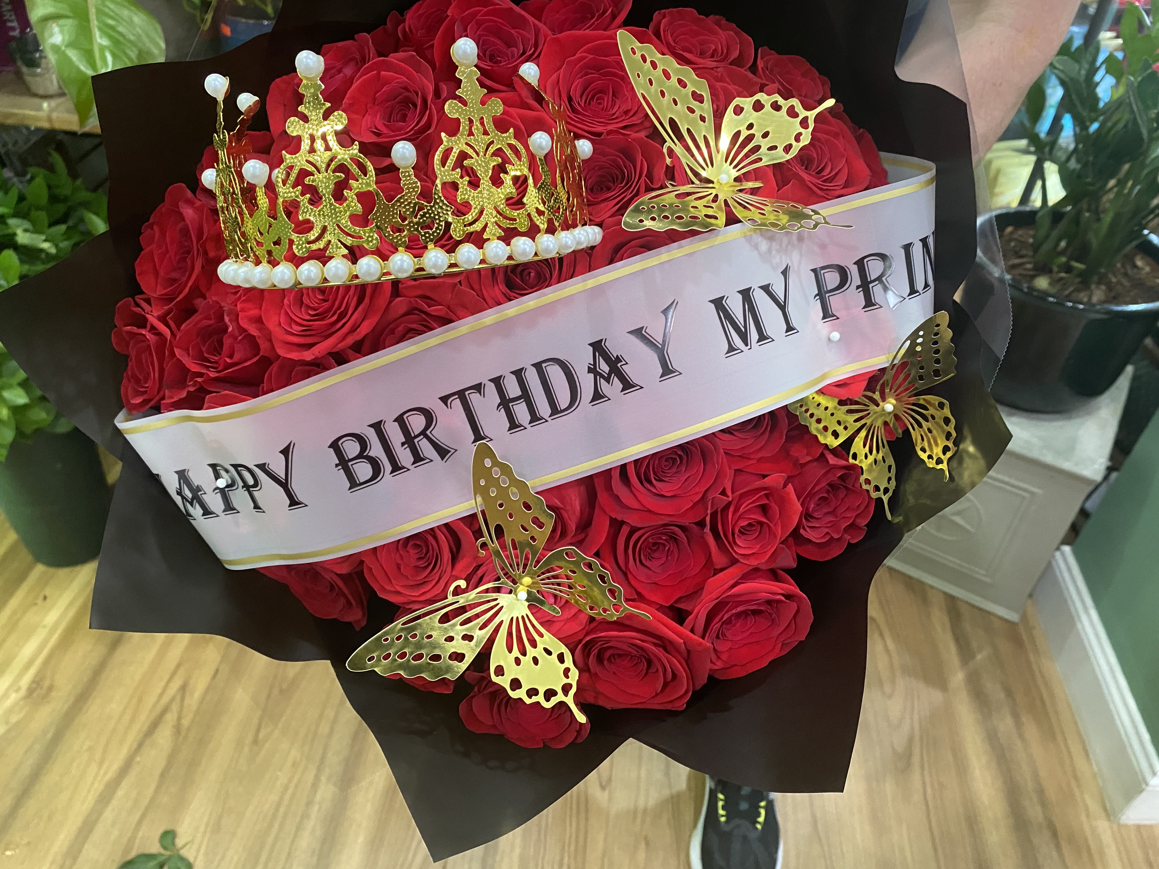Birthday Hand Bouquet - A bold cluster of red roses designed to make the birthday person feel crowned and celebrated - this birthday hand bouquet pairs classic romance with dramatic flair. The red roses and black wrapping paper create a striking contrast that reads both festive and sophisticated, perfect for a birthday gift, a milestone celebration, or an anniversary with a playful edge.  Tied with a pale birthday sash across the front, the bouquet is finished with a decorative gold crown topped with pearl-like details and several gold butterfly picks nestled among the blooms. The rich red roses are tightly arranged for a dense, luxurious look; black wrapping paper gives modern structure while the yellow and white accents add tiny flashes of brightness against the deep palette.  Give this to someone you want to make feel truly seen on their special day - the crown and sash announce the occasion while the roses deliver the emotion. Hand-deliver or have it sent as a standout birthday surprise; keep blooms in fresh water and away from direct heat to extend vase life.