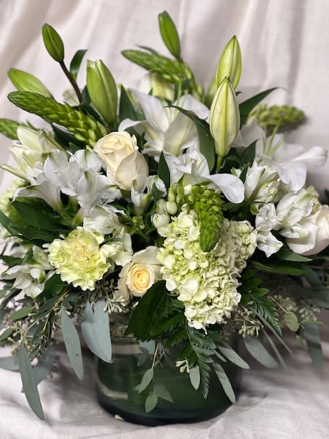 Green Delight - A serene, layered arrangement of white and soft green blooms, Green Delight is a white flower arrangement perfect for Mother's Day, an anniversary, or to say "I'm thinking of you" with grace. The low glass vase and the calming palette make this an especially fitting choice for sympathy or funeral services, while also elegant enough for weddings, thank-you gifts, or a refined housewarming present.  White Asiatic lilies and unopened lily buds rise above a dense cluster of cream roses, pale green-white hydrangeas, white chrysanthemums, alstroemeria, and carnations to create a full, textured silhouette. Soft gray-green foliage spills around the base, giving the design subtle contrast and a gentle, natural movement. The arrangement is presented in a clear, low glass vase that highlights the layered stems and compact, rounded shape.  