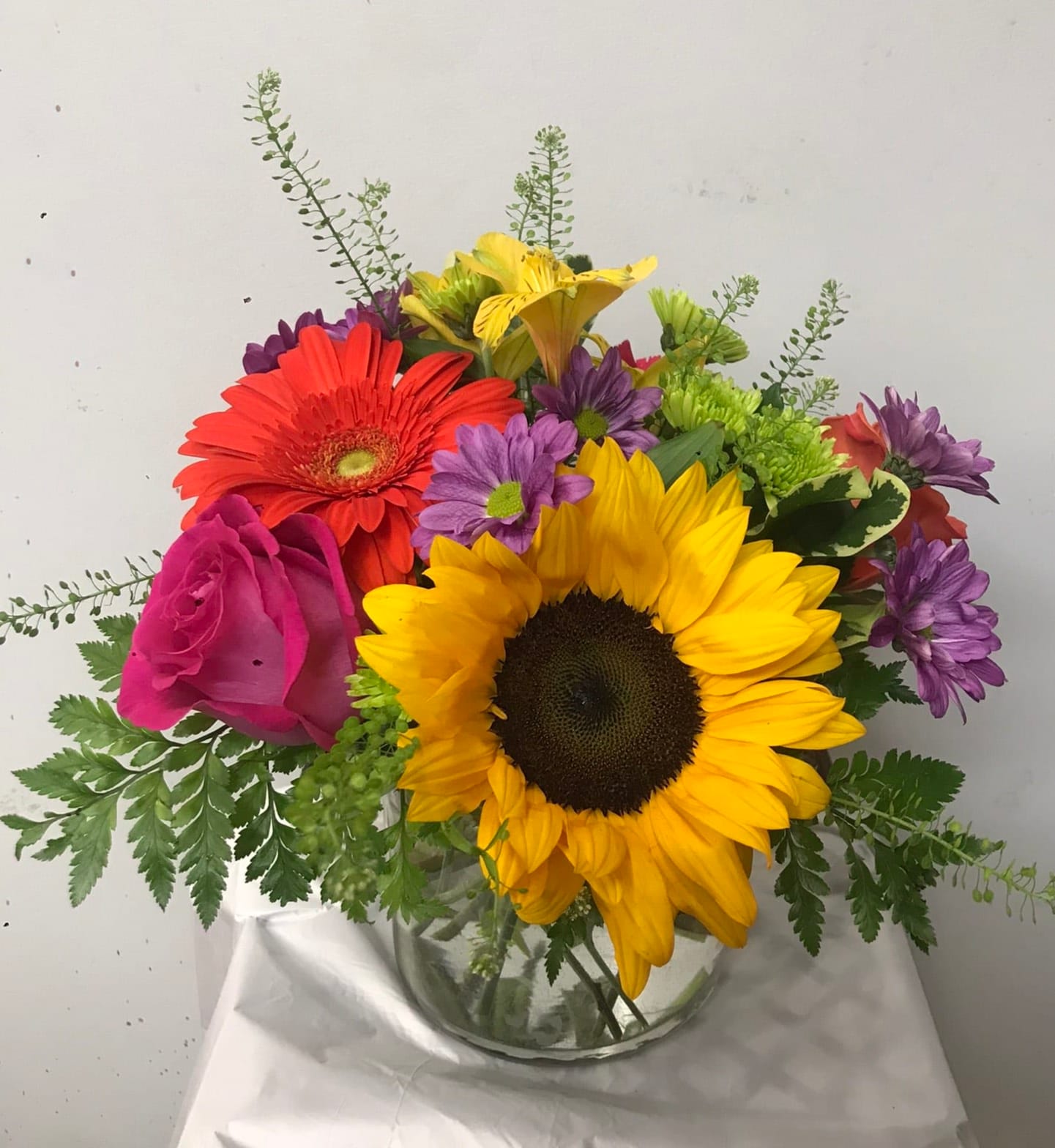 Mixed bouquet with sunflowers, roses, gerbera daisies, and purple blooms in a glass vase