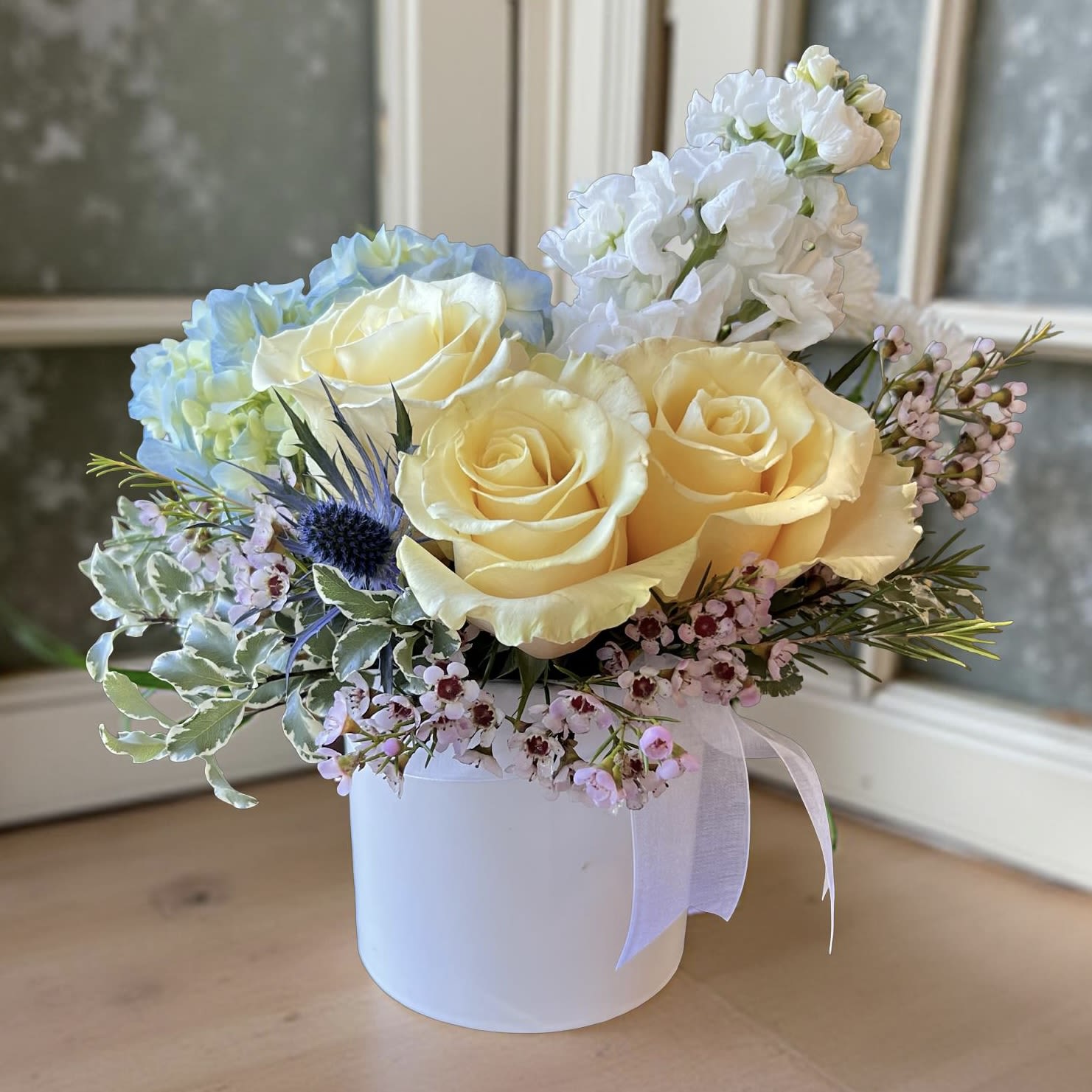 Yellow roses and blue hydrangea in a white hat box with pink filler flowers.