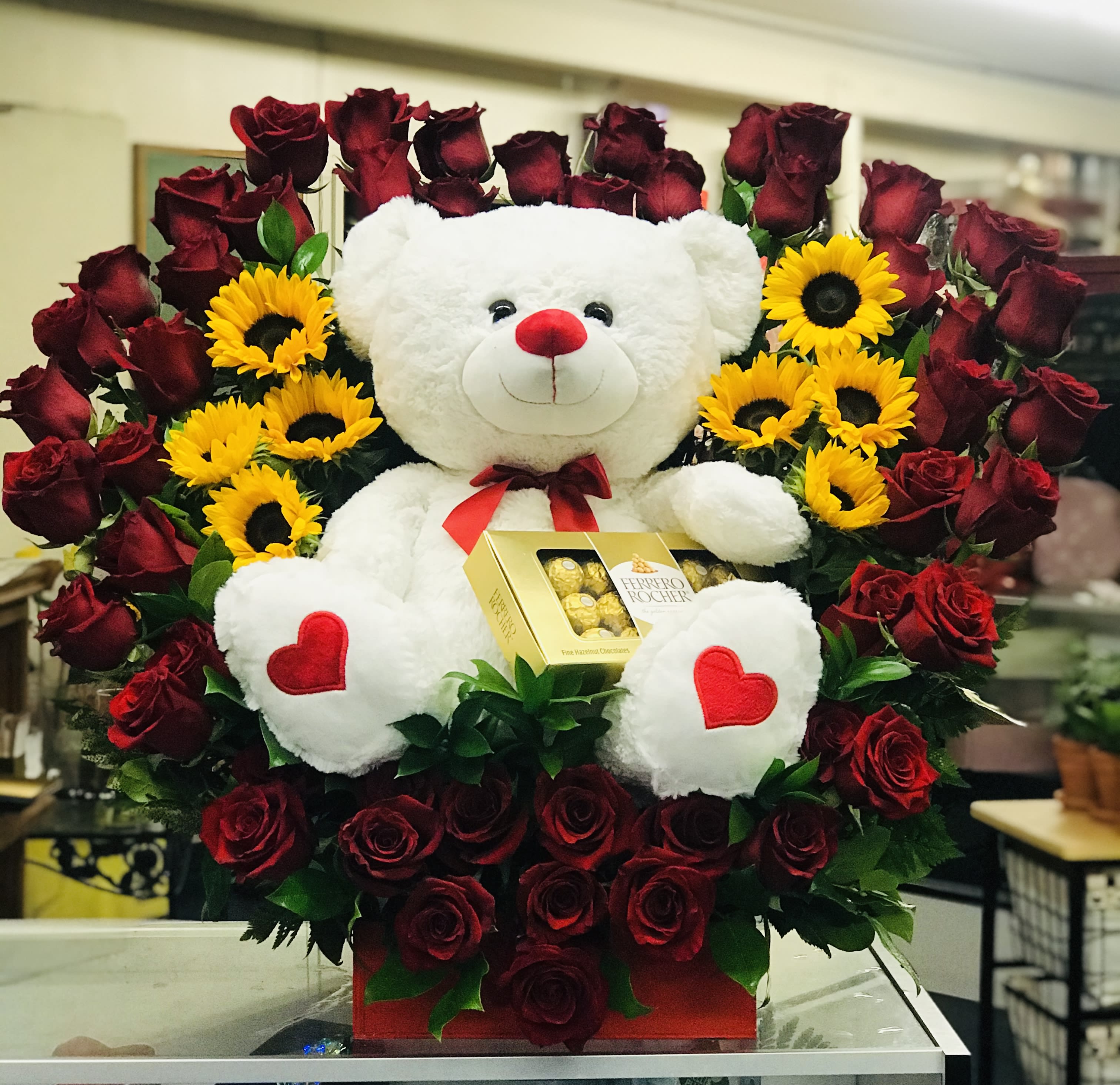 Cupids love cloud - This arrangement has about 55-60 roses in a heart shape arch where a beautiful bear seats over the middle holding a box of chocolates. Also seats near sunflowers and ruscus greenery to complete the seat to this big arrangement.