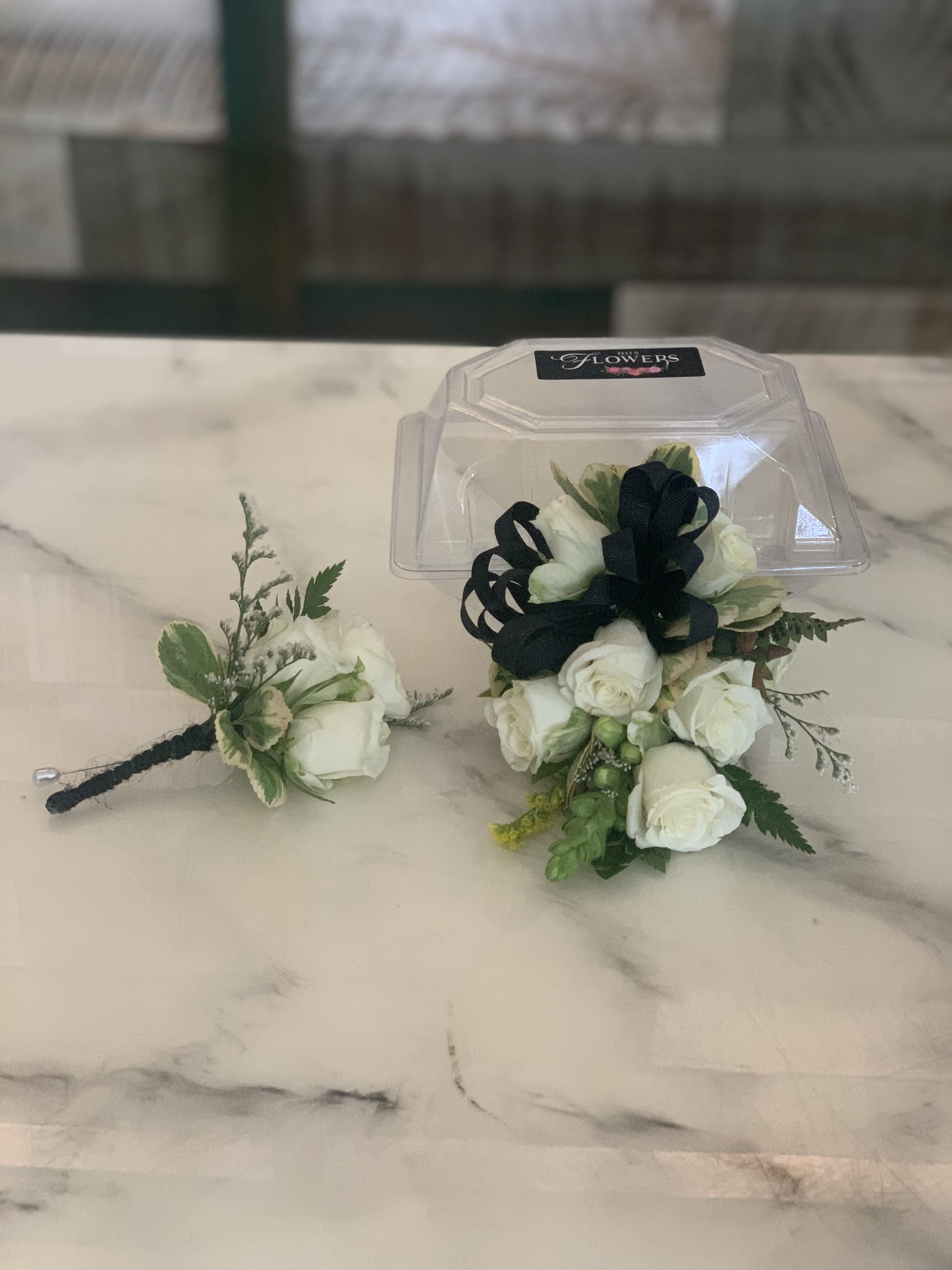 White rose boutonniere with black ribbon and greenery beside a clear plastic case