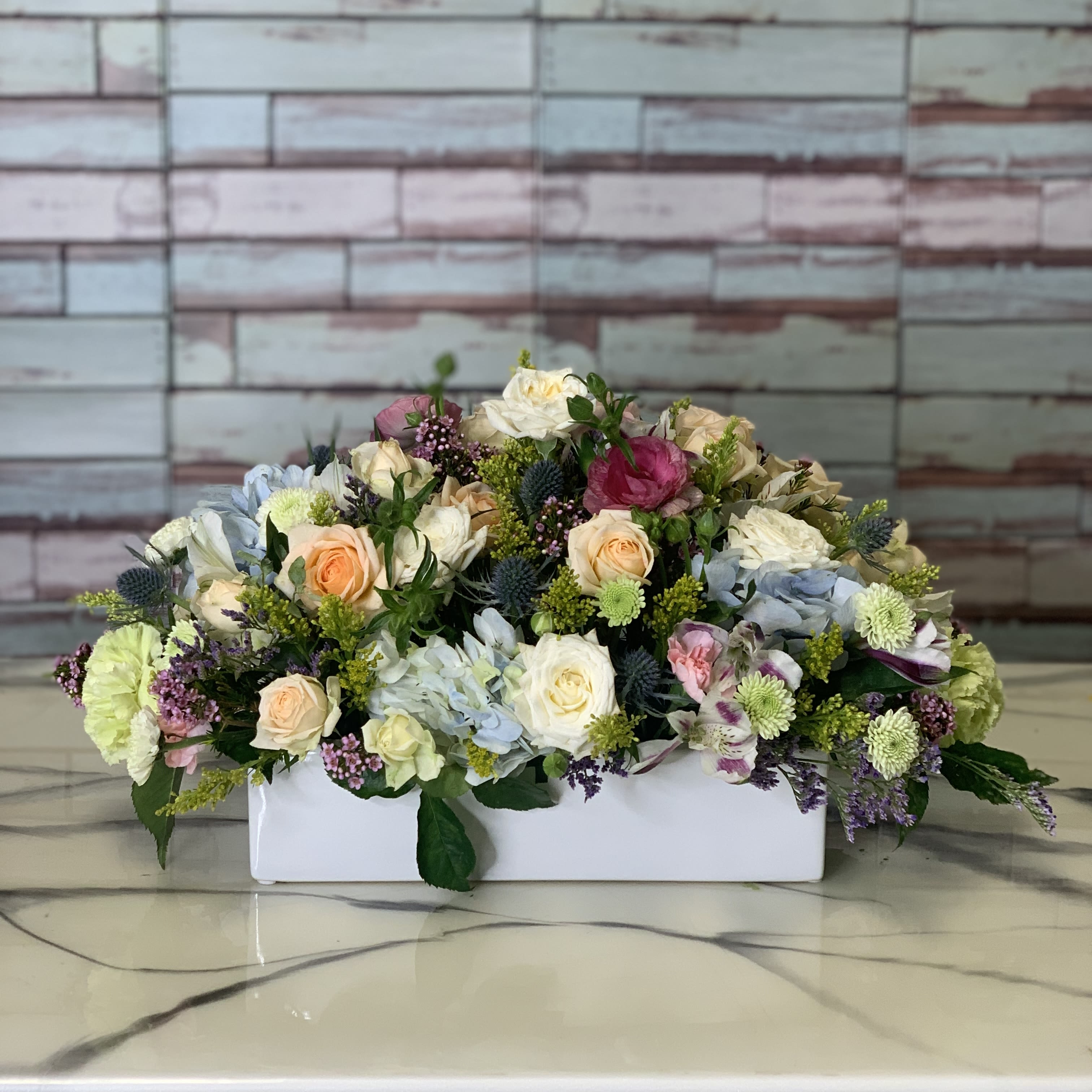Country Chic Centerpiece - A country chic centerpiece features seasonal blooms to include hydrangea, roses, sprays, assorted mums, mini gerberas, and greenery with fill. Arranged low and compact in a white ceramic. Perfect to take to a dinner party or send to someone just because!