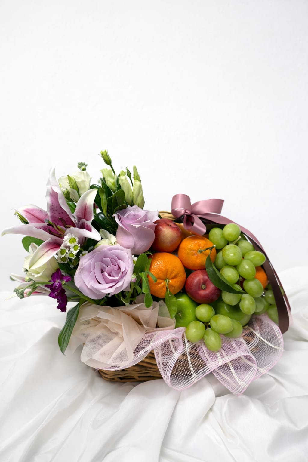 Basket arrangement with lilies, roses, and fruit topped with a pink ribbon