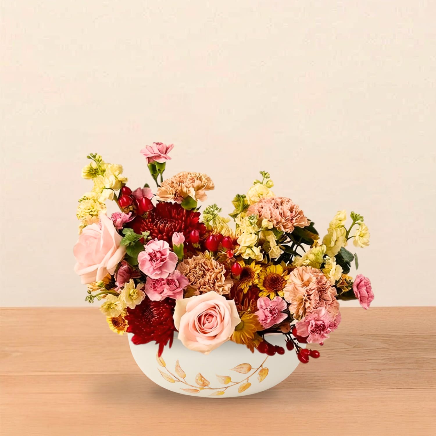 Bountiful Centerpiece - A warm and abundant fall centerpiece, handcrafted in our signature Bountiful style.  Celebrate the season with a lush, full centerpiece inspired by harvest hues and cozy gatherings. The Bountiful Bowl is designed in an elegant white vessel adorned with a golden leaf motif, a nod to autumn’s beauty.  Each arrangement features a rich mix of roses, mums, carnations, berries, and seasonal blooms in shades of peach, amber, red, and blush. Its low, wide shape makes it perfect for dining tables, coffee tables, or sideboards, offering warmth and elegance without blocking conversation. Arranged in a keepsake ceramic bowl (8.25" wide x 3" tall).  Please note: The photo shown is a reference for the color palette and overall style. Every arrangement is handcrafted and unique, with flower varieties and fullness varying slightly based on seasonal availability and the selected upgrade.