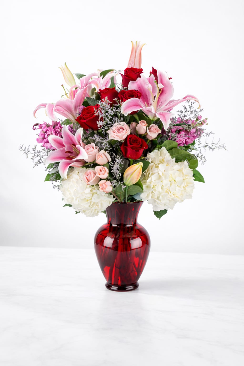 Bouquet of pink lilies, red roses, and white hydrangeas in a red glass vase