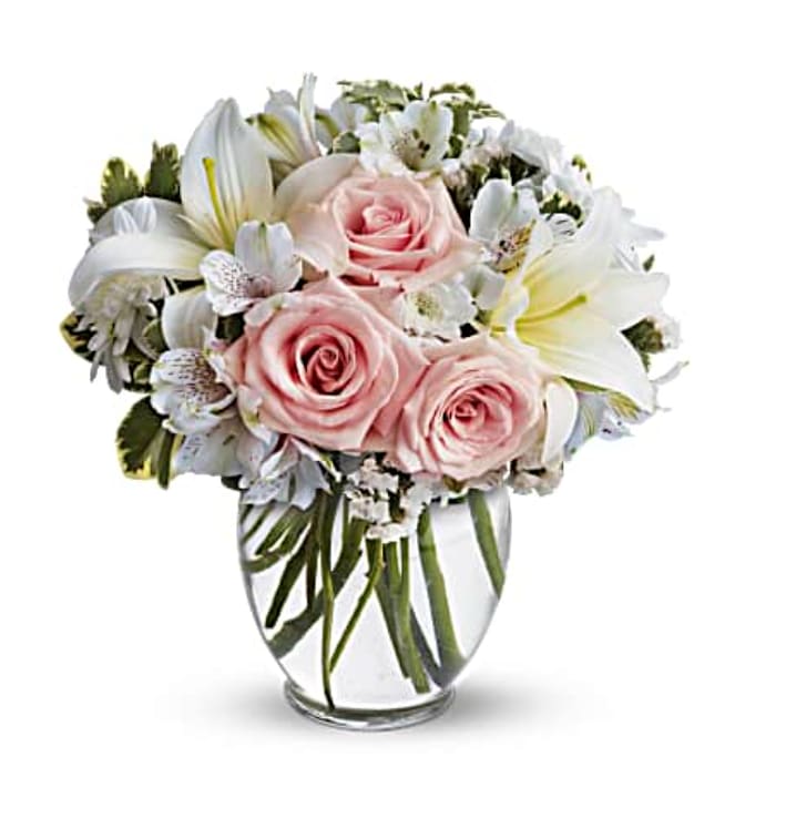 Whites and Pinks ( TelaFlora ) - light pink roses, white asiatic lilies, white alstroemeria and white cushion spray chrysanthemums are mixed with white statice and variegated pittosporum in a clear, rounded vase.