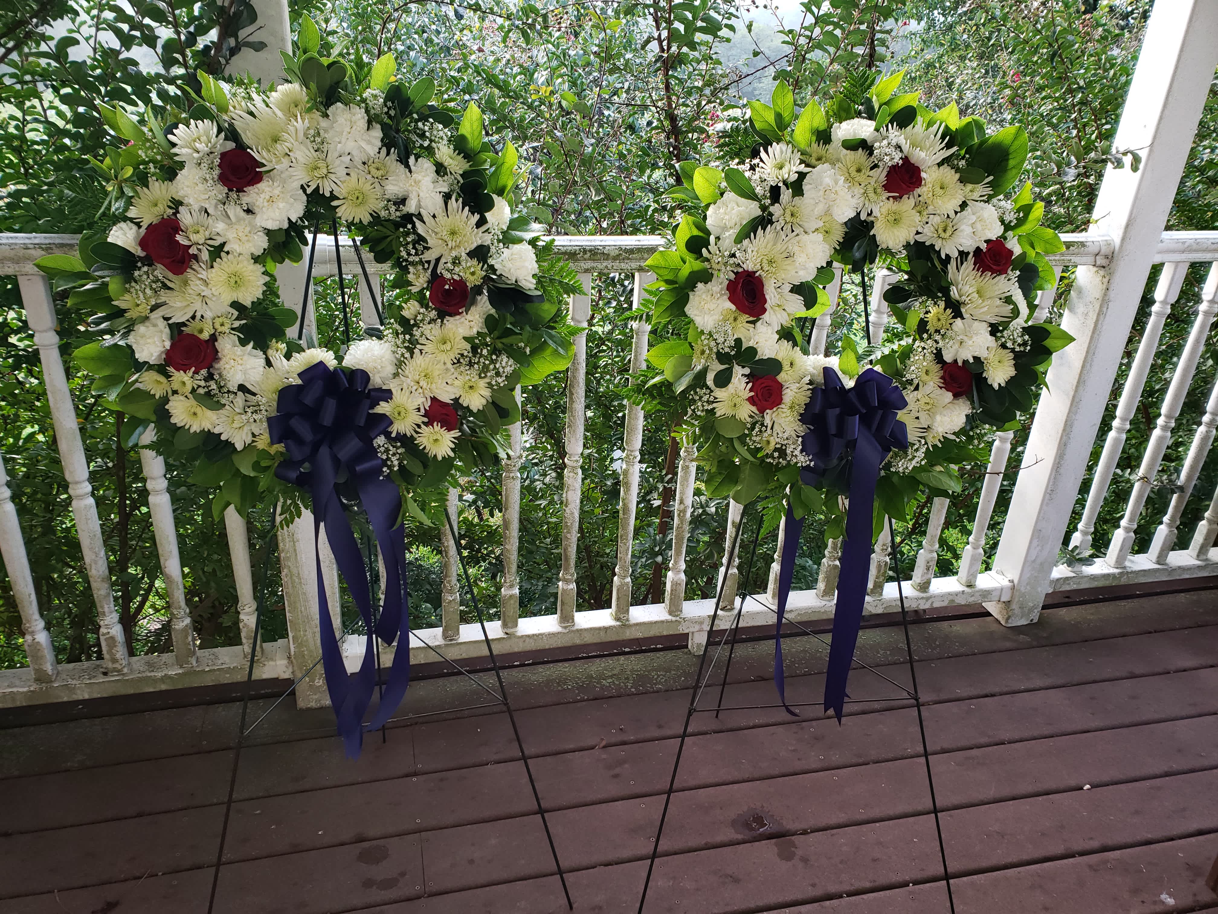 Two floral wreaths on stands with white flowers, red roses, and navy ribbons