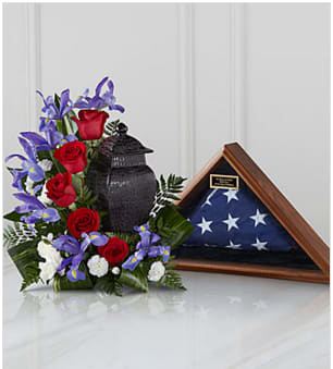Red roses and purple flowers arranged beside a folded American flag in a display case