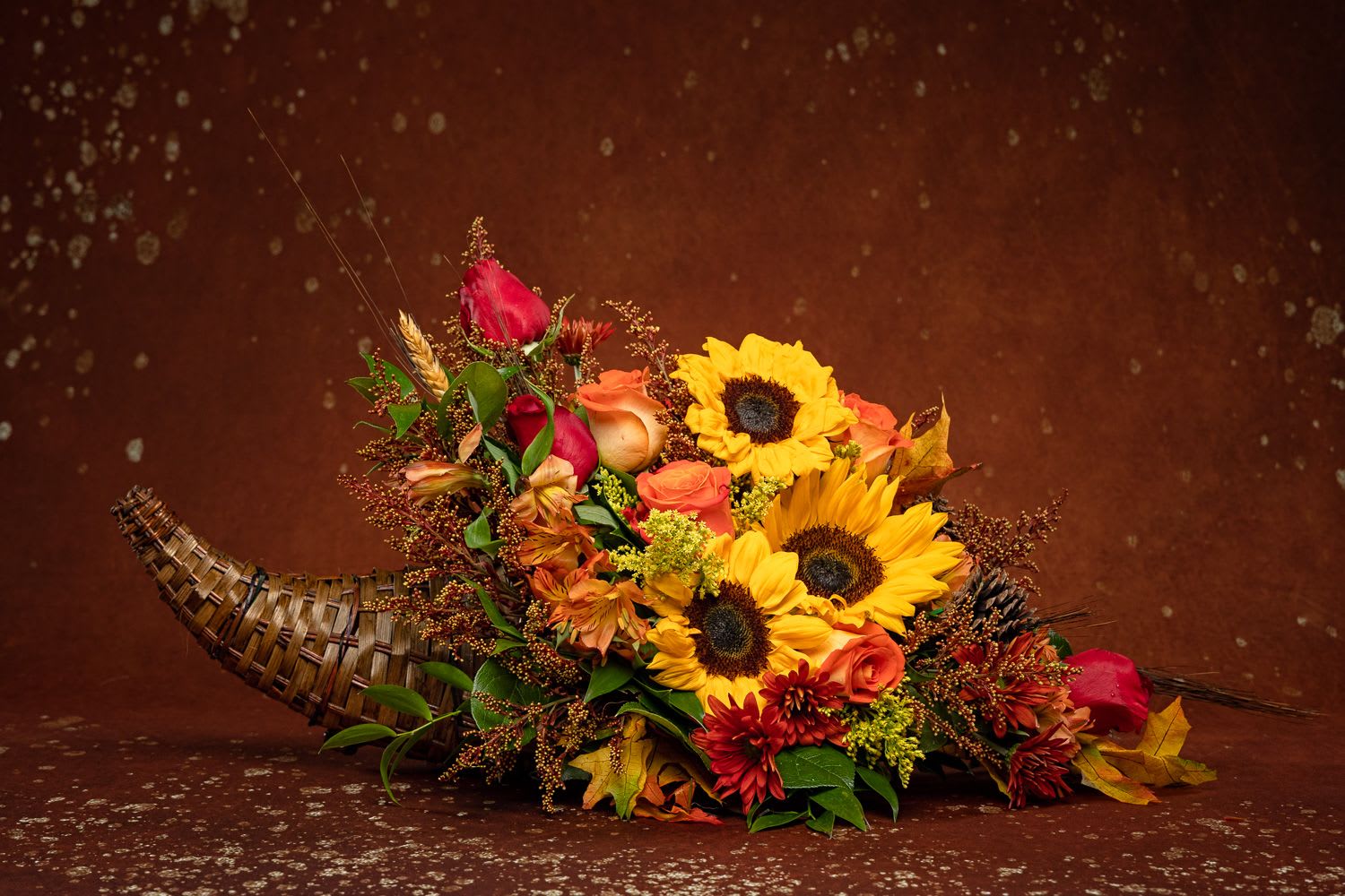 Cornucopia  - *Classic Cornucopia* - This month our Charity in Bloom Arrangement will support feeding our locals who are less fortunate. We will be using 10% of this arrangement to provide food to people in our local community, That will be without food on thanksgiving day. Thank you for your support to this great cause.  *Celebrate the season of Thanksgiving with this traditional symbol of autumnal abundance. The perfect Thanksgiving table decoration, this beautiful cornucopia is rendered as a horn-shaped wicker basket filled with fall flowers and harvest wheat. Also, ideal for the kitchen counter, coffee table or entryway. 	•	Gorgeous sunflowers and lush orange roses are accented with unique red daisies, yellow cushion mums, parvifolia eucalyptus and magnolia leaves in a wicker cornucopia-shaped basket. 	• harvest wheat are comforting fall touches. 	•	Orientation: One-Sided  