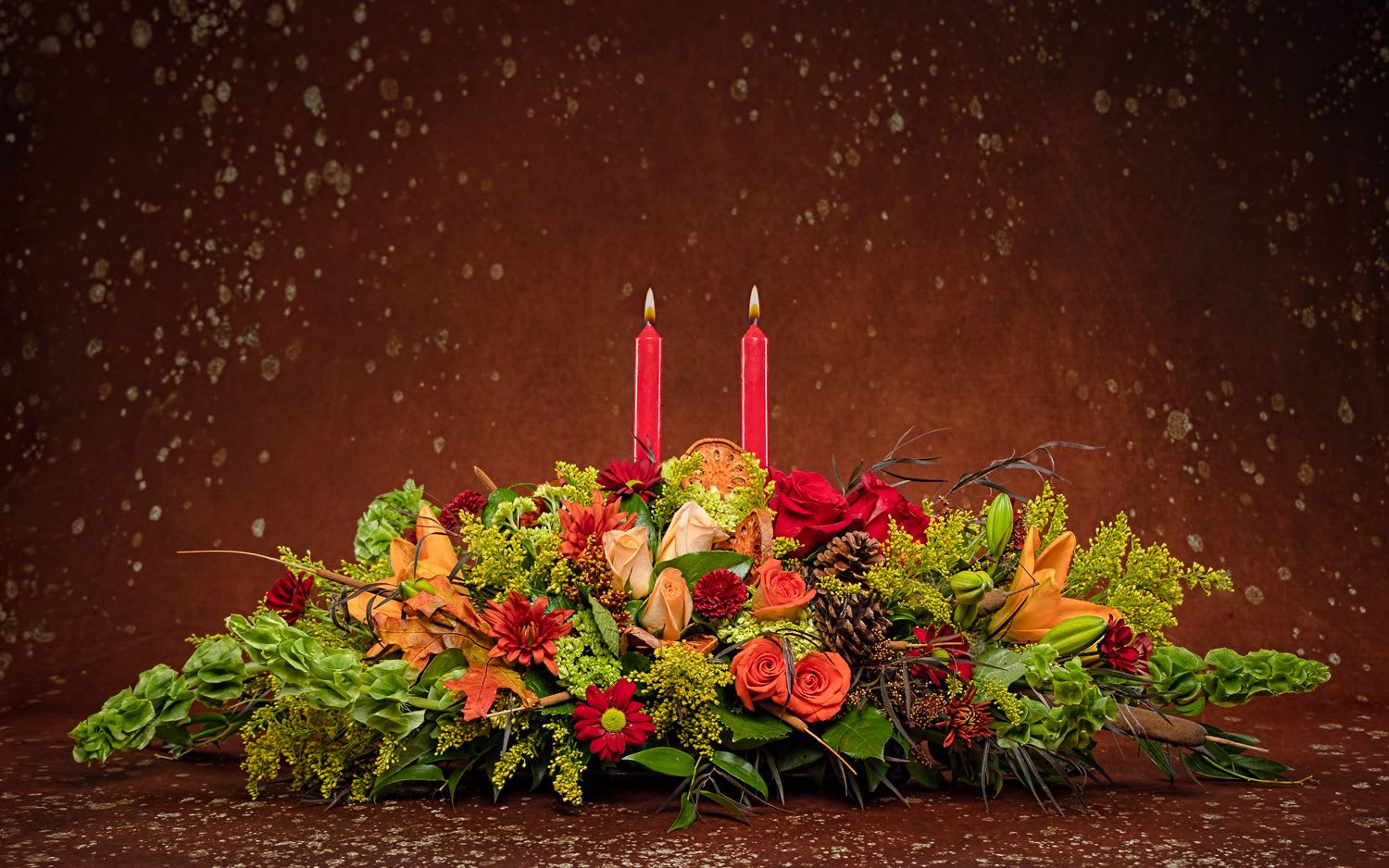 Thanksgiving Centerpiece  - Graciously gather your family around the warm glow and classic autumn colors of this stunning Thanksgiving centerpiece. A lush variety of burnt orange blooms are gathered under a pair of tall, graceful candles - perfect for decorating your Thanksgiving dinner table, entryway or hearth. Fall flowers include orange roses, orange asiatic lilies, green Bells of Ireland dark orange alstroemeria, bronze button mums, yellow cushion mums, rust cushion mums, green oregonia and green huckleberry. Accented with oak leaves and two Red taper candles. Orientation: All-Around