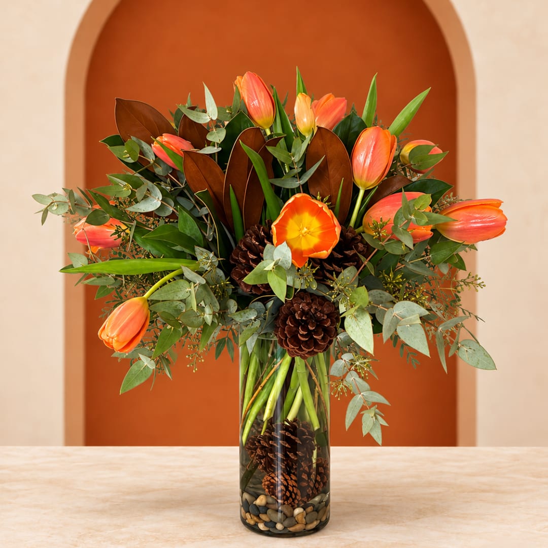 Orange tulips arranged in a glass vase with pinecones and greenery.