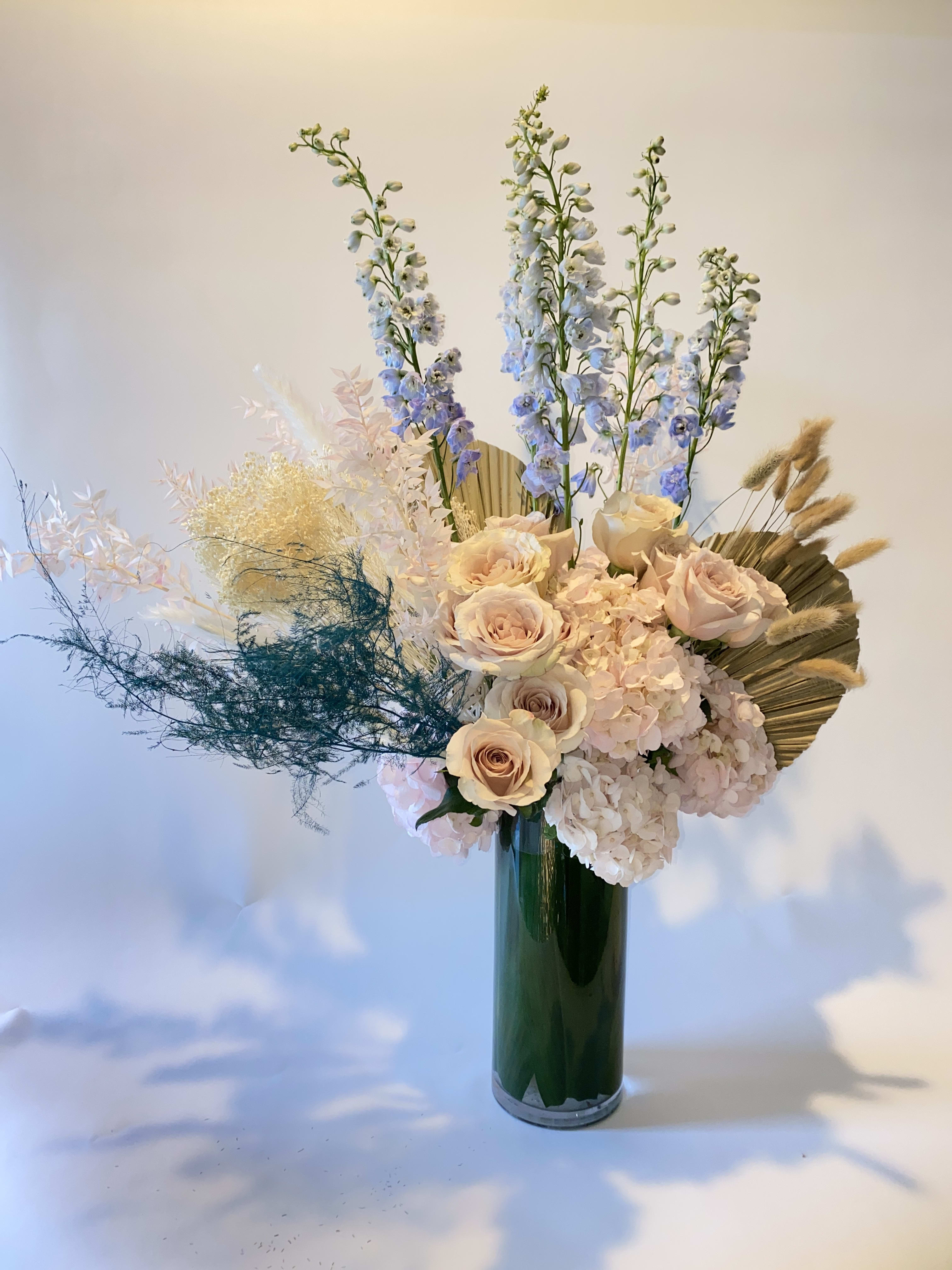 Tall floral arrangement with pale roses, hydrangeas, and blue delphinium in a green vase
