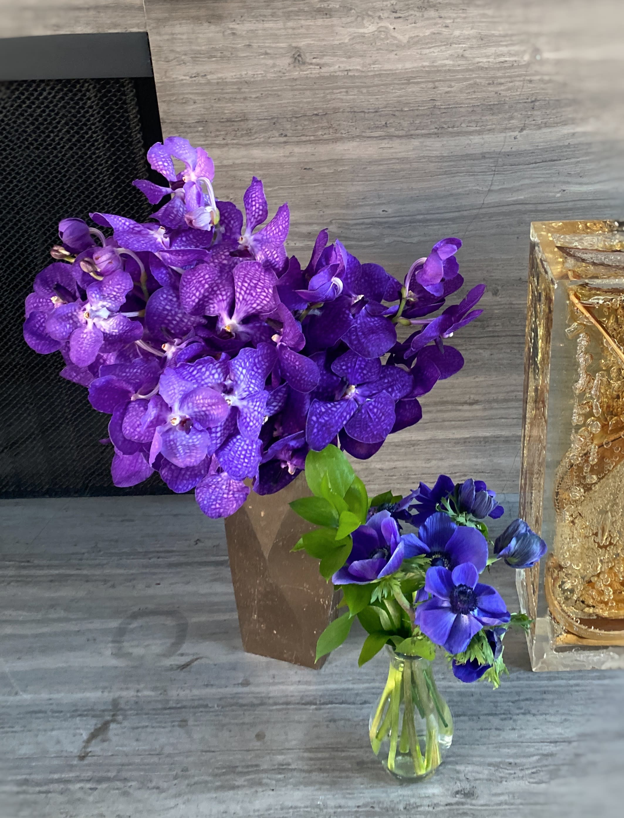 Purple orchid arrangement in a brown vase beside a small blue flower bouquet in a glass bottle