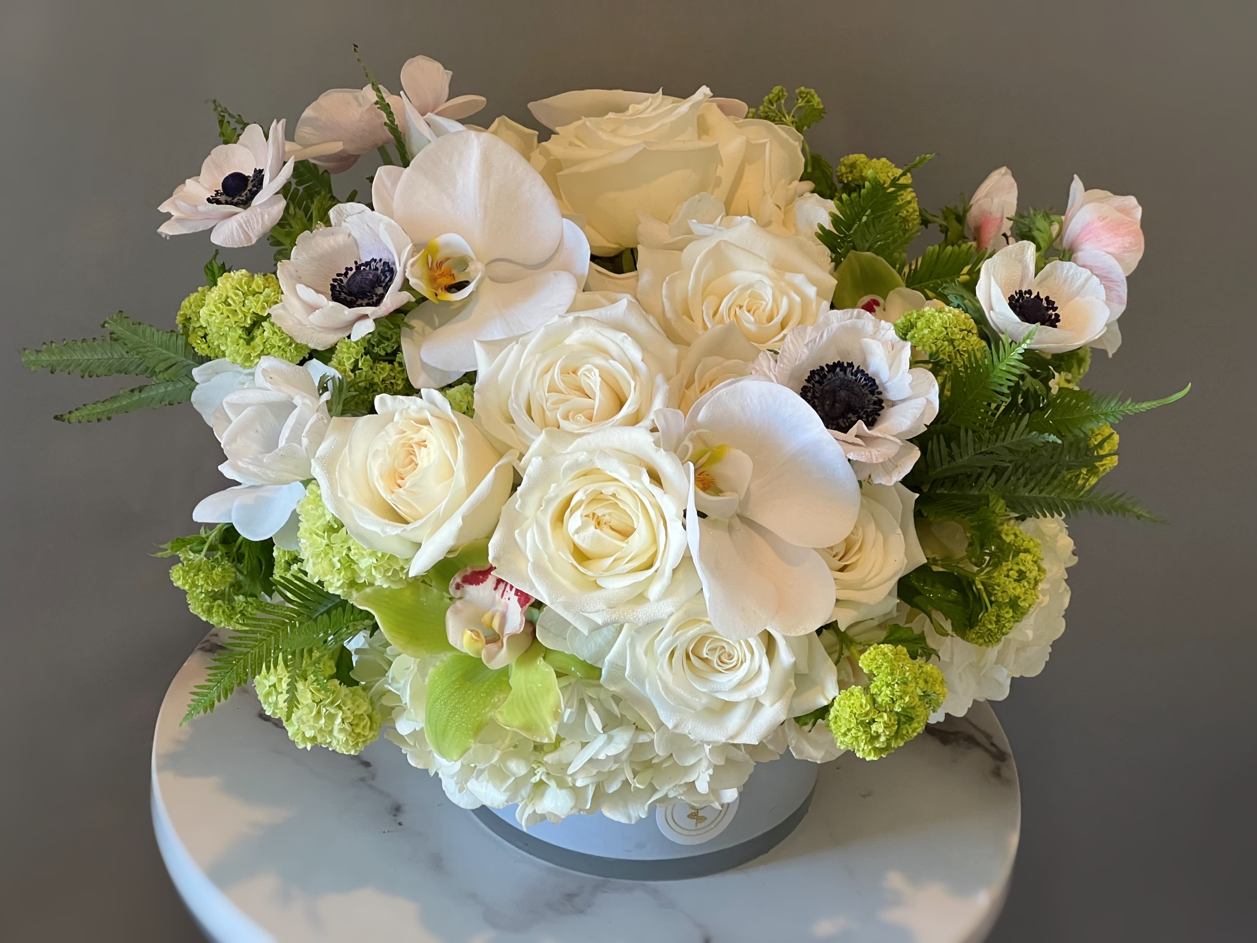 White roses and orchids arranged in a round box with green accents