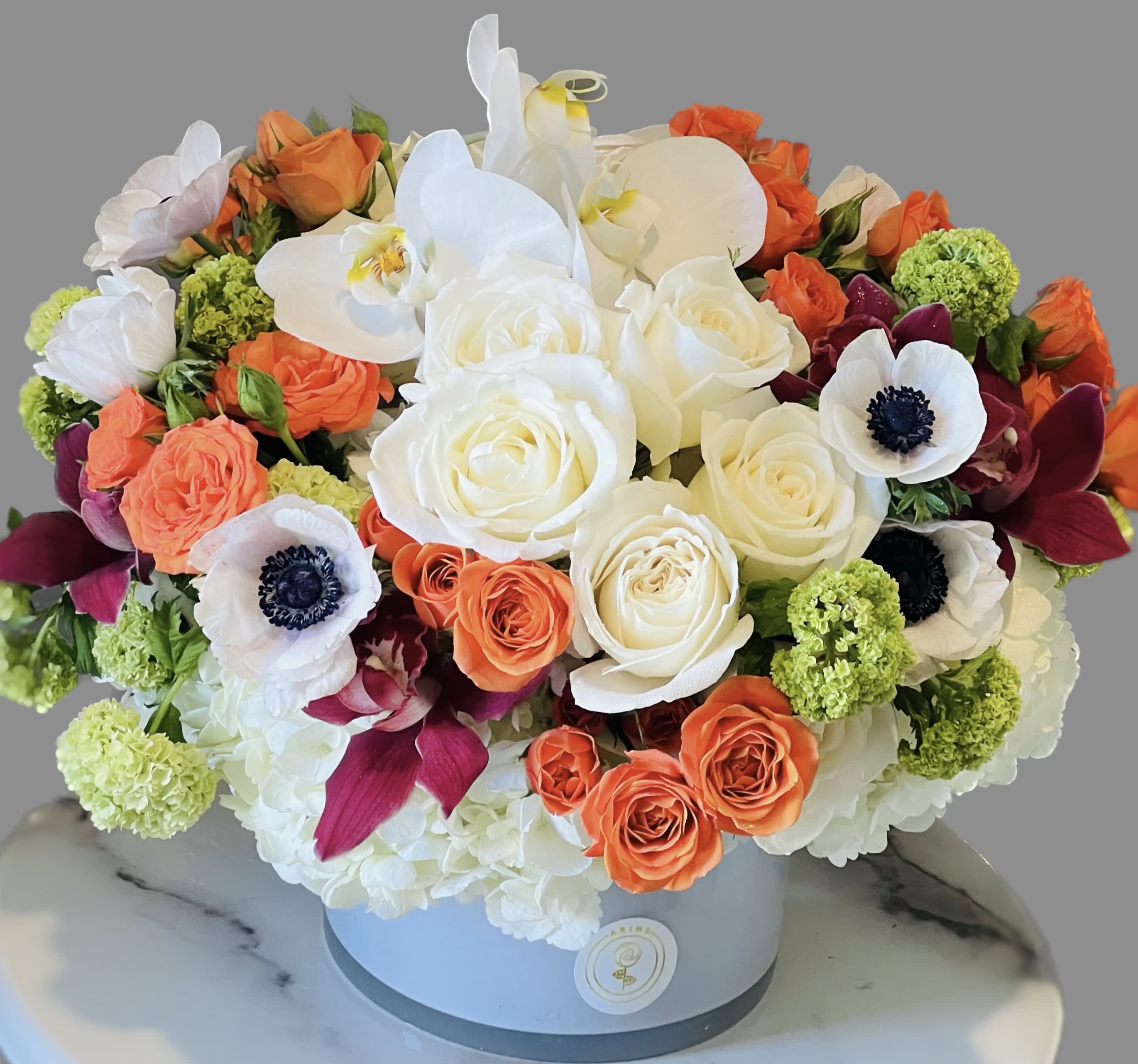 Round bouquet of white orchids, roses, and orange blooms in a hatbox.
