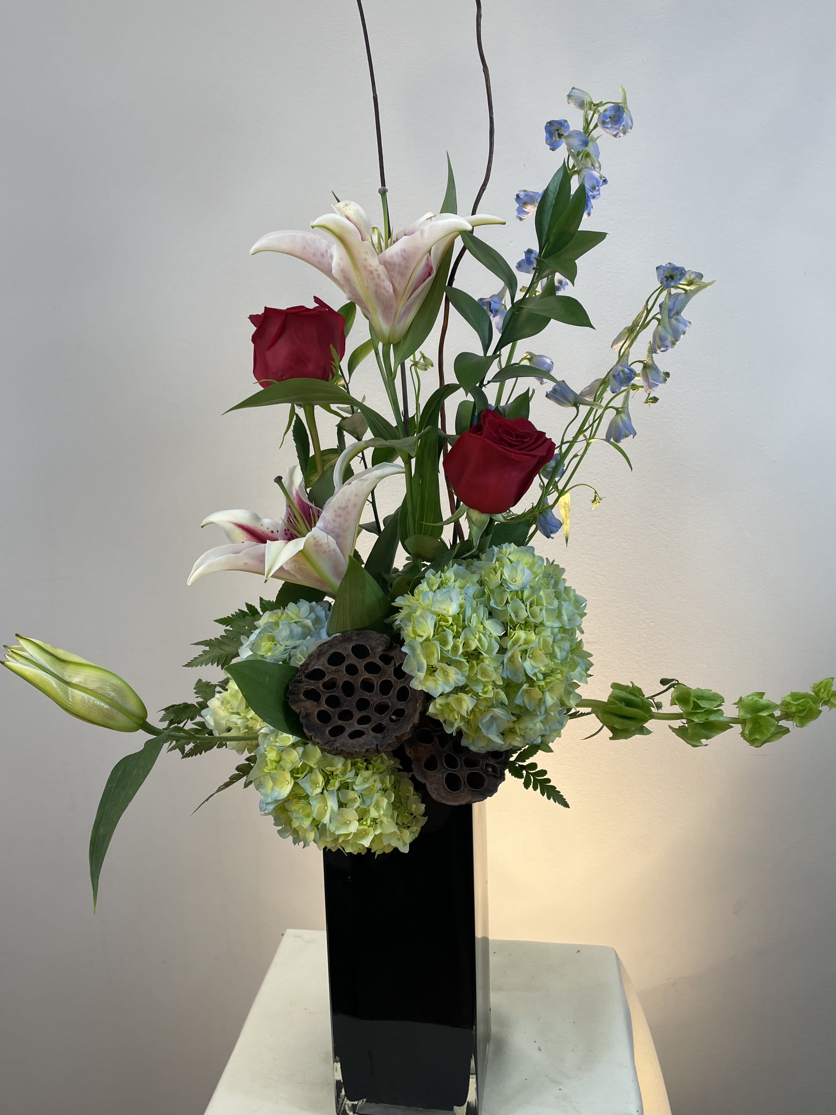 Tall floral arrangement with red roses, lilies, and blue flowers in a black vase