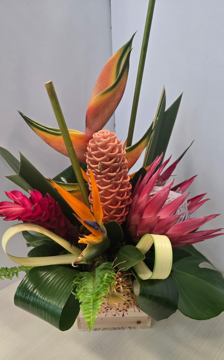 Tropical Flame Fusion by Art and Flowers - THIS IS A SPECIAL ORDER DESIGN.  We will source the needed topicals and deliver as soon as possible. It might be a 3 to 4 day wait due to availability  of the flowers. We will keep you informed as to delivery day.  Please order this design in advance of delivery date.  This bold tropical arrangement showcases a striking mix of texture, form, and vibrant color. A sculptural peach ginger cone rises at the center, framed by dramatic heliconia blooms with warm orange and yellow tones. A vivid bird of paradise adds a burst of fiery contrast, while a rich pink king protea brings depth and intensity.  Glossy green leaves are artfully looped and layered, creating movement and a modern, architectural feel. Delicate fern accents soften the composition, while the natural wooden container grounds the design with an organic touch.   