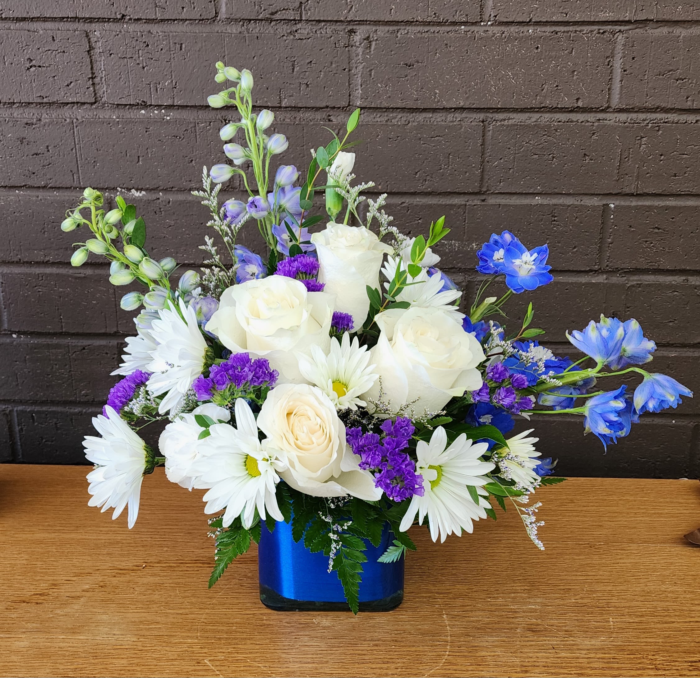 Blue in the Blue Ridge - Crisp white roses paired with cheerful white daisy-like blooms and vivid purple filler create a fresh, airy bouquet that feels like a clear mountain morning - perfect as a birthday bouquet, a thank-you gift, or a general celebration arrangement. The asymmetrical, lively shape with taller stems rising above the center gives it movement and an effortless, joyful presence that will make the person you're thinking of feel genuinely noticed.  Bright blue accent flowers pop against the white and purple palette and arrive arranged in a striking blue glass vase that deepens the color story and brightens any tabletop. The design is full with side sprays for a natural silhouette and works well for everyday gifting; its mixed textures and cool tones suit recipients who love a modern, cheerful look. Care: trim stems and refresh water every few days to keep blooms looking their best.