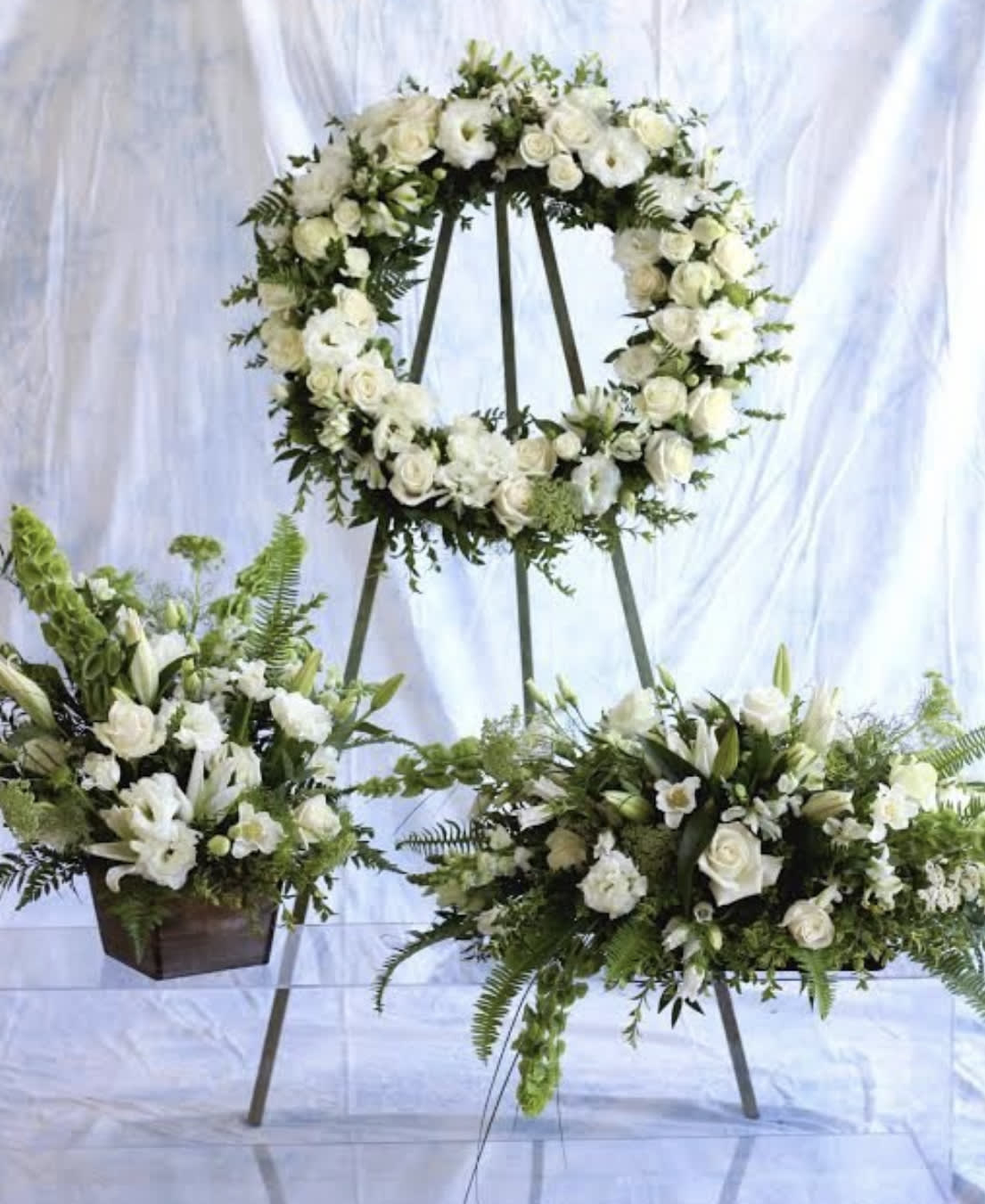 White wreath, spray, and matching floral arrangement on stands with green accents for a sympathy display.