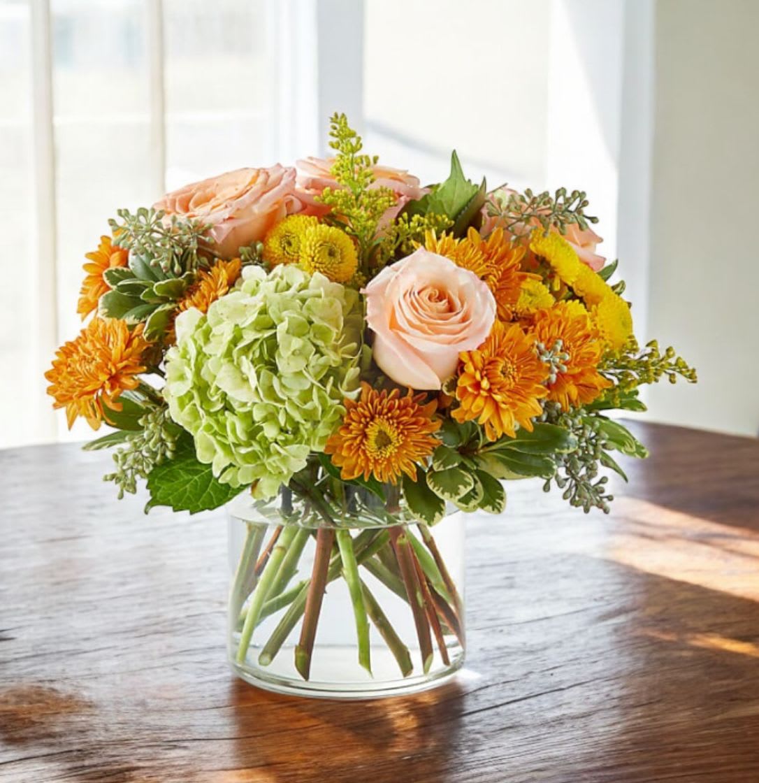 Round arrangement of peach roses, green hydrangea, and orange mums in a clear glass vase