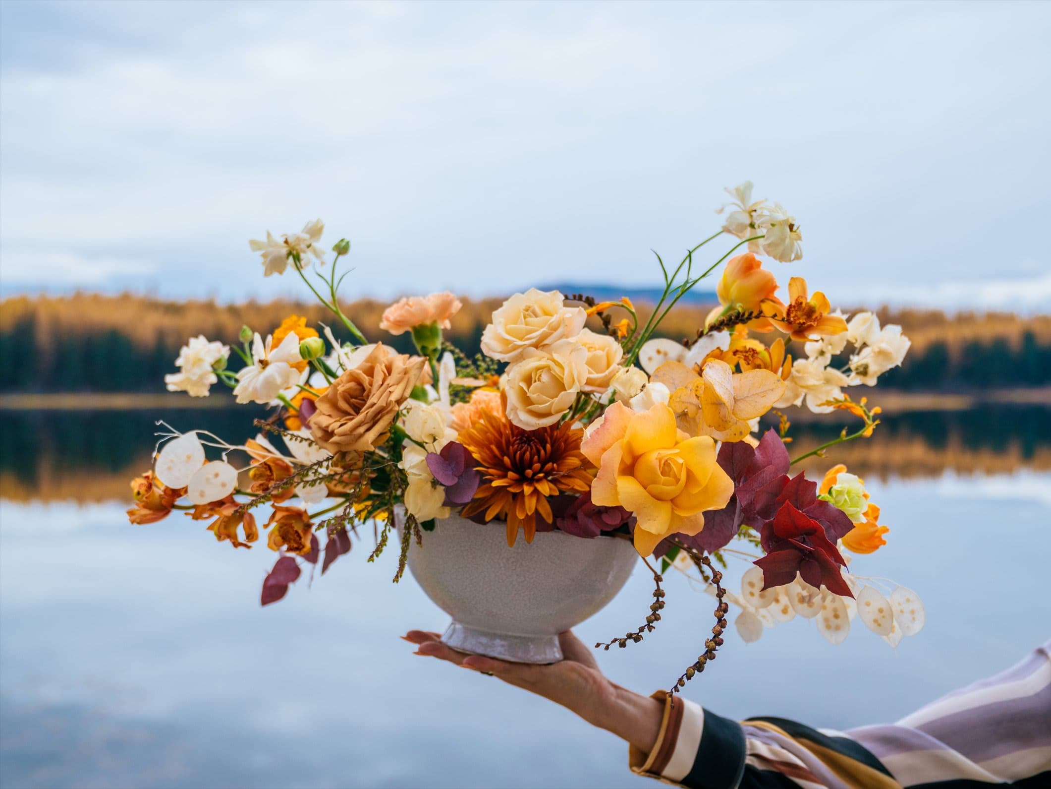 Low centerpiece of yellow, peach, and cream flowers in a white pedestal bowl held by a hand.