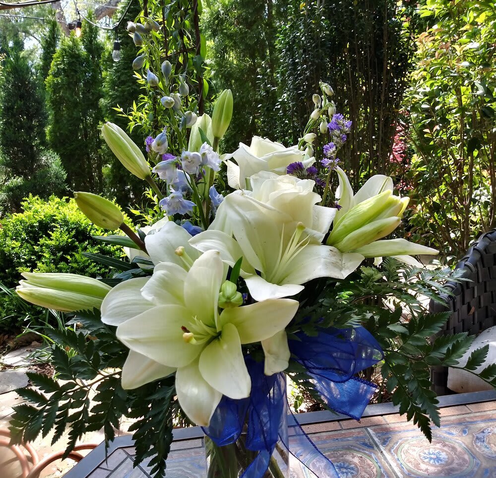 White lily and rose arrangement with blue ribbon and purple accents in a clear vase on an outdoor table