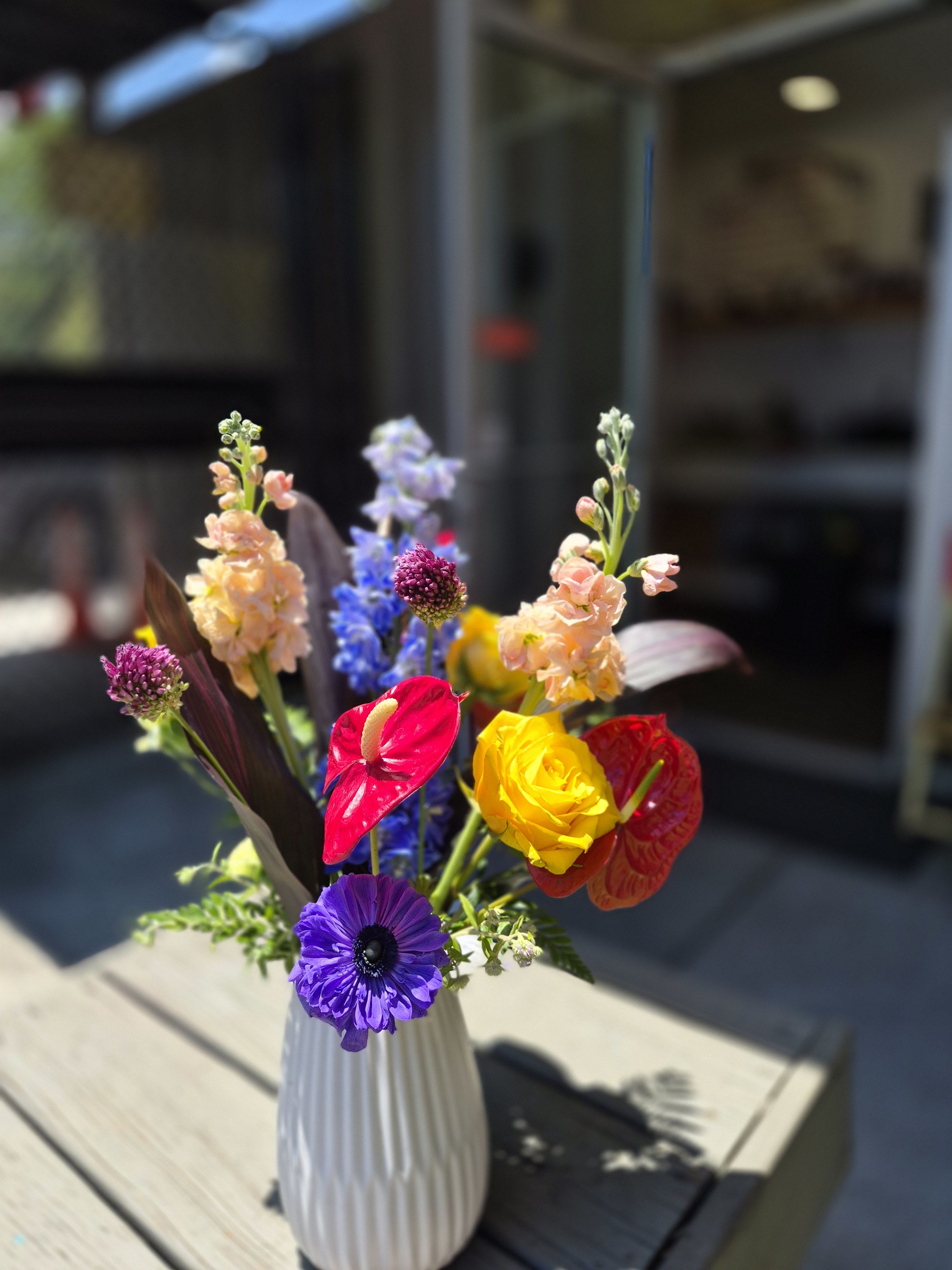 Adams Park Posy -  The Adams Park Posy is a vibrant celebration of one of Southwest Atlanta's most beloved historic communities. Inspired by the neighborhood's picturesque landscape, enduring legacy, and welcoming spirit, this bouquet reflects the beauty found where history and nature flourish together.  Bold red anthuriums symbolize the strength and resilience of the Adams Park community, while delicate pink lisianthus adds a touch of Southern grace and hospitality. Golden yellow roses evoke the warmth of neighbors gathering beneath the community's mature tree canopy, and striking blue delphinium mirrors the tranquil waters and open skies surrounding the neighborhood's historic park. Accented with lush greenery, each stem is thoughtfully arranged to create a design that is both sophisticated and joyful.  The Adams Park Posy is more than a bouquet; it is a floral tribute to one of Atlanta's landmark communities, where vibrant colors, rich history, and enduring neighborhood pride continue to bloom.  