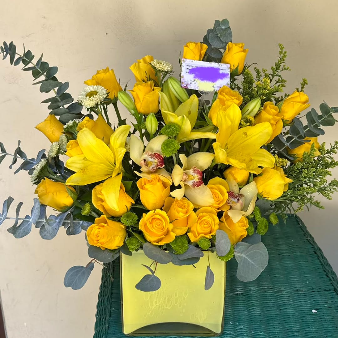 Yellow roses and lilies arranged in a square vase with eucalyptus