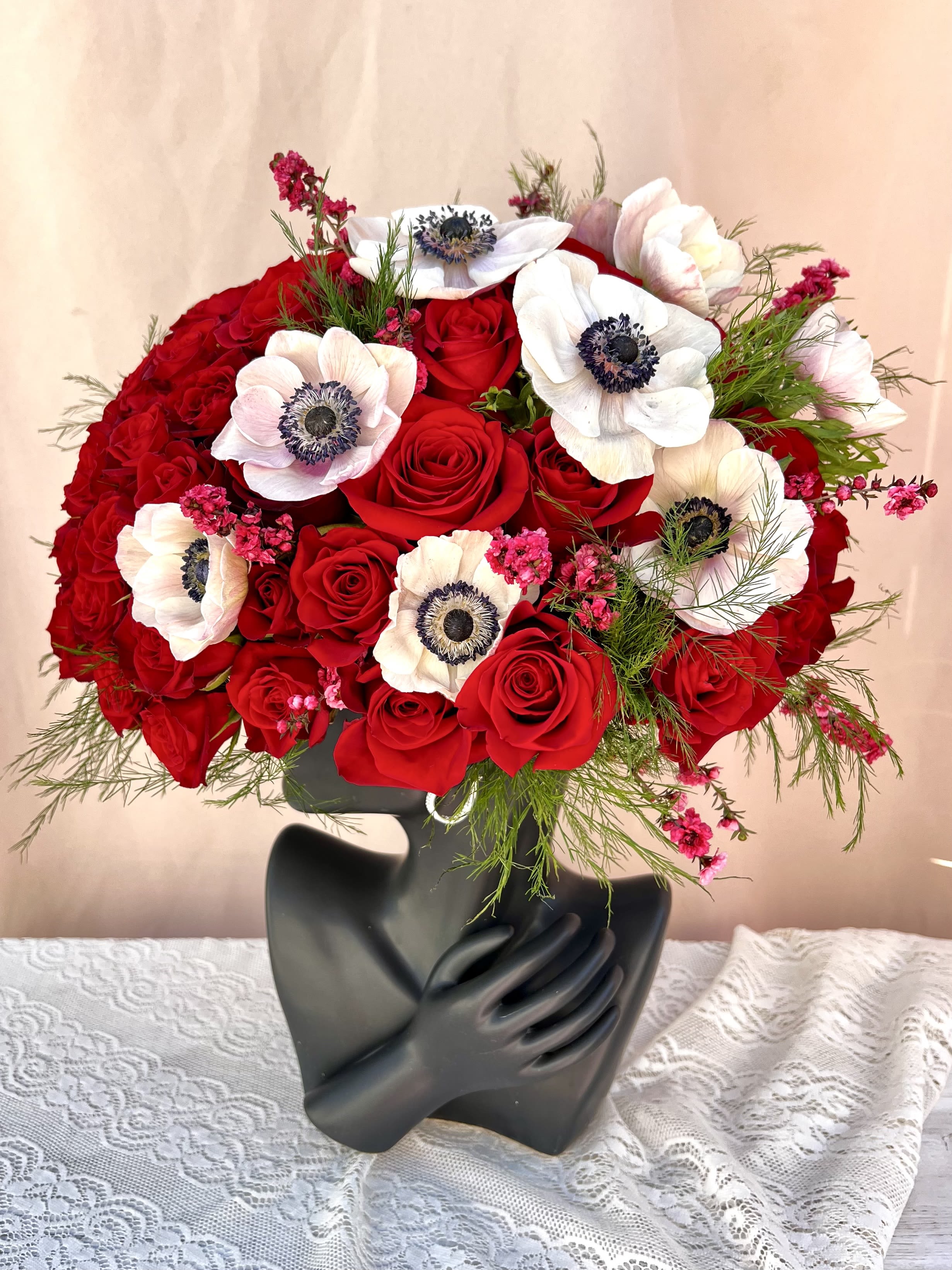 Red rose and white anemone arrangement in a black sculptural face vase on a lace-covered table.
