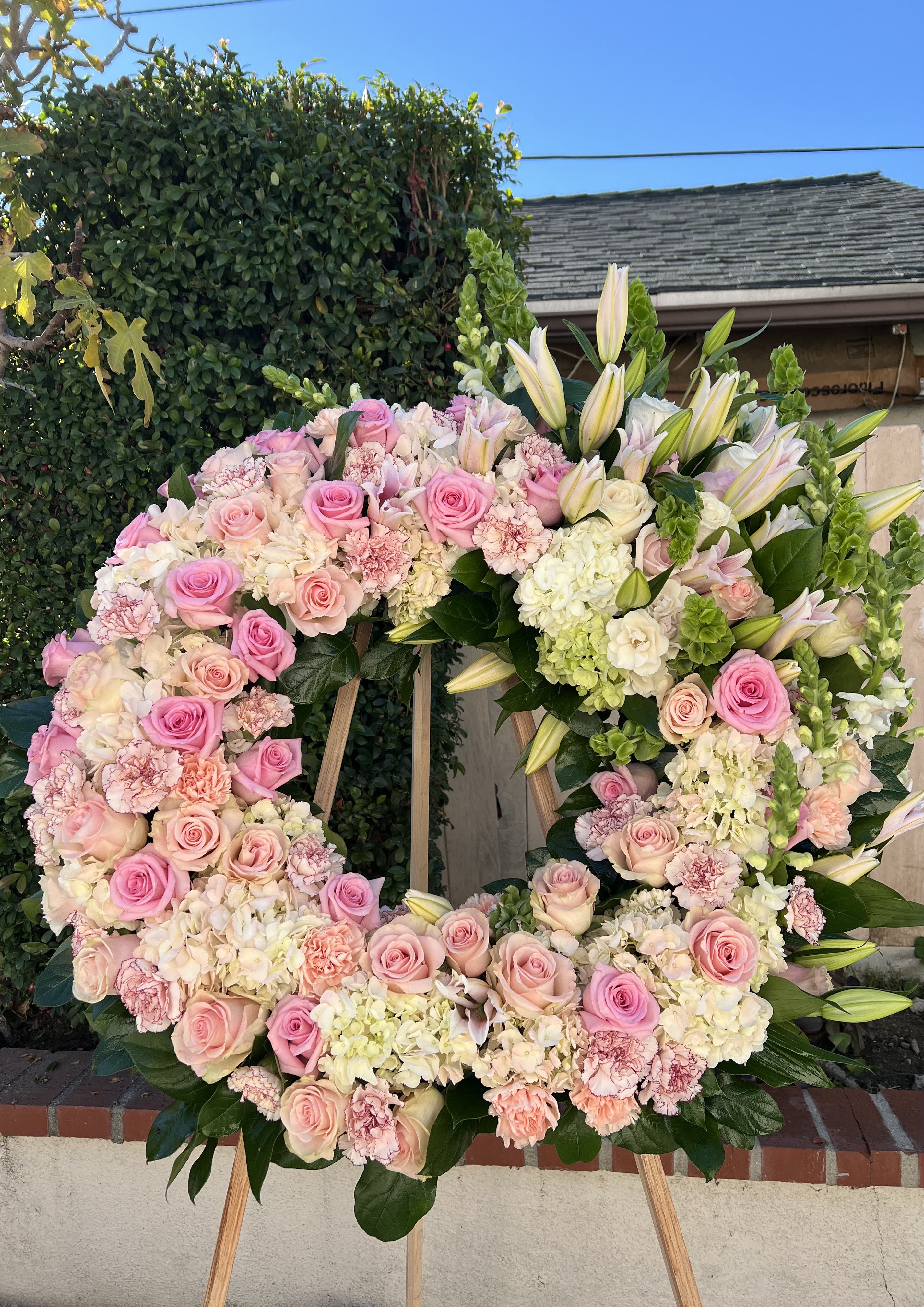 Large standing wreath of pink roses, white hydrangeas, carnations, and lilies on a wooden easel.