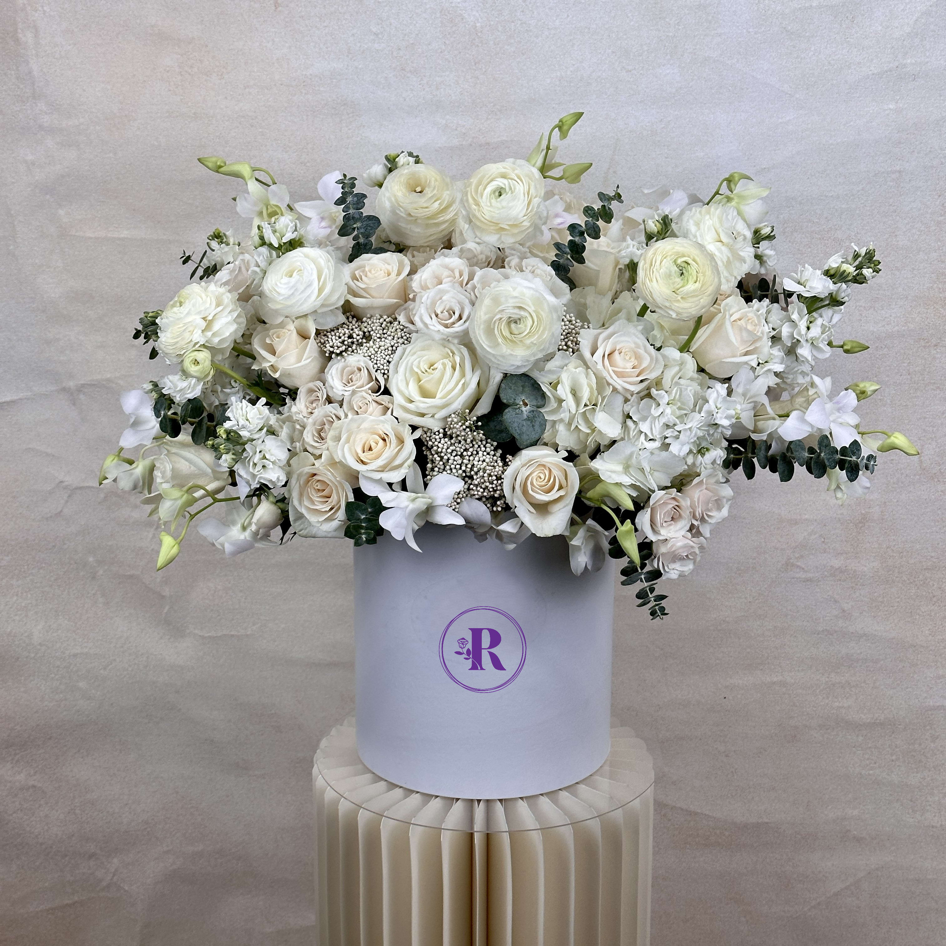 Large white and cream rose and ranunculus arrangement in a round white hatbox on a pleated pedestal.