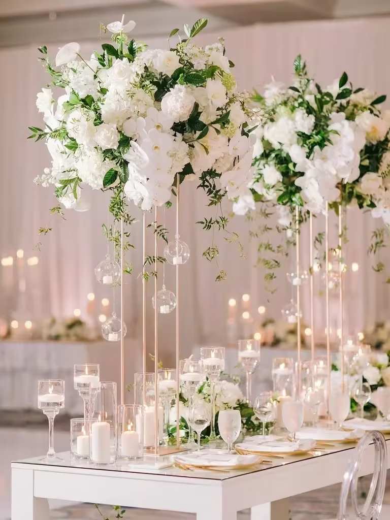 Tall white wedding centerpieces with orchids and roses on gold stands above a candlelit reception table